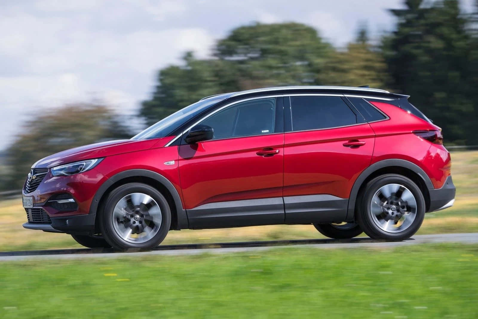 Opel Grandland X 桌布 壁紙 — 一輛鮮紅色的SUV在草叢路邊疾馳，背景有模糊的綠樹和陰天。