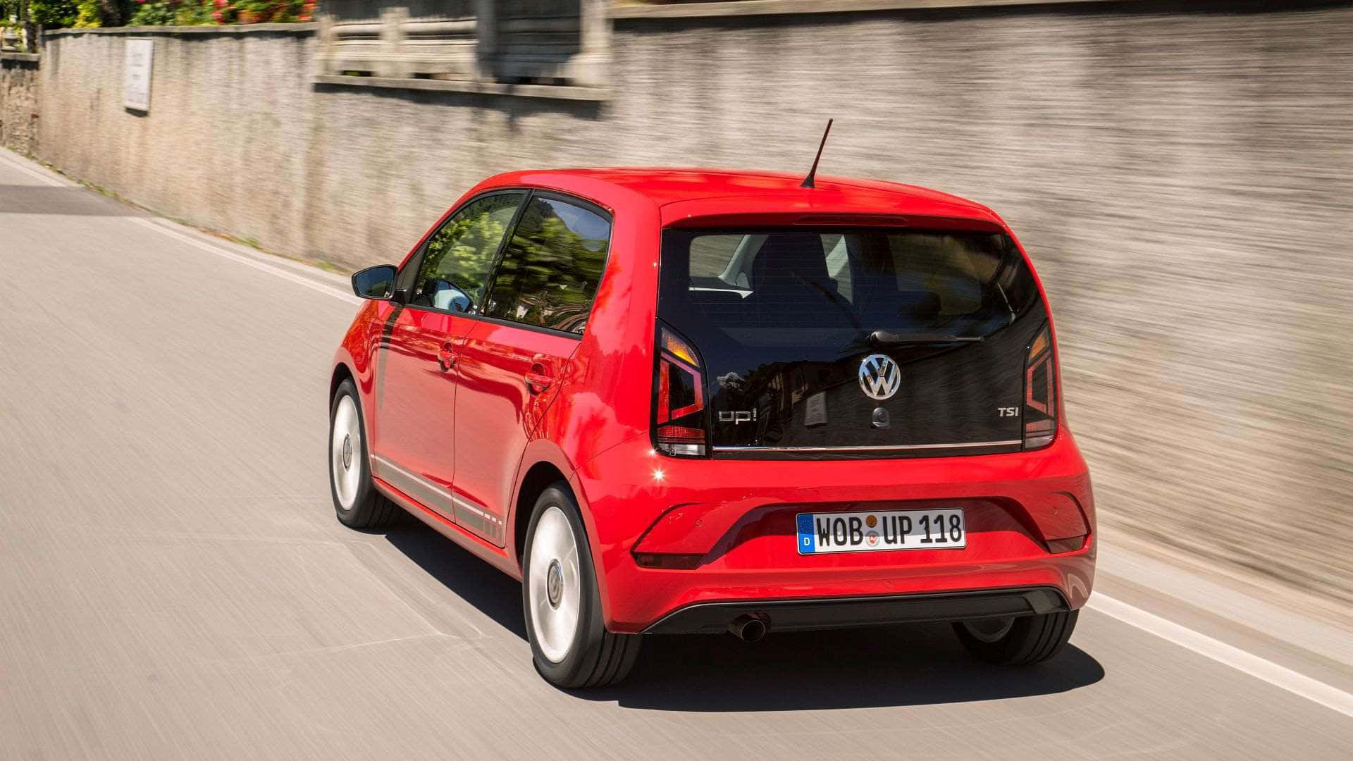  壁紙 — 一輛充滿活力的紅色Volkswagen up! TSI掀背車在陽光明媚的道路上行駛，背景是模糊的石牆。