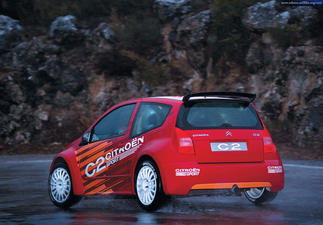 Citroen C2 Super 1600 桌布 壁紙 — 一輛充滿活力的紅色雪鐵龍 C2 Sport 拉力賽車在濕滑的瀝青路面上濺起水花，背景是崎嶇的岩石懸崖。
