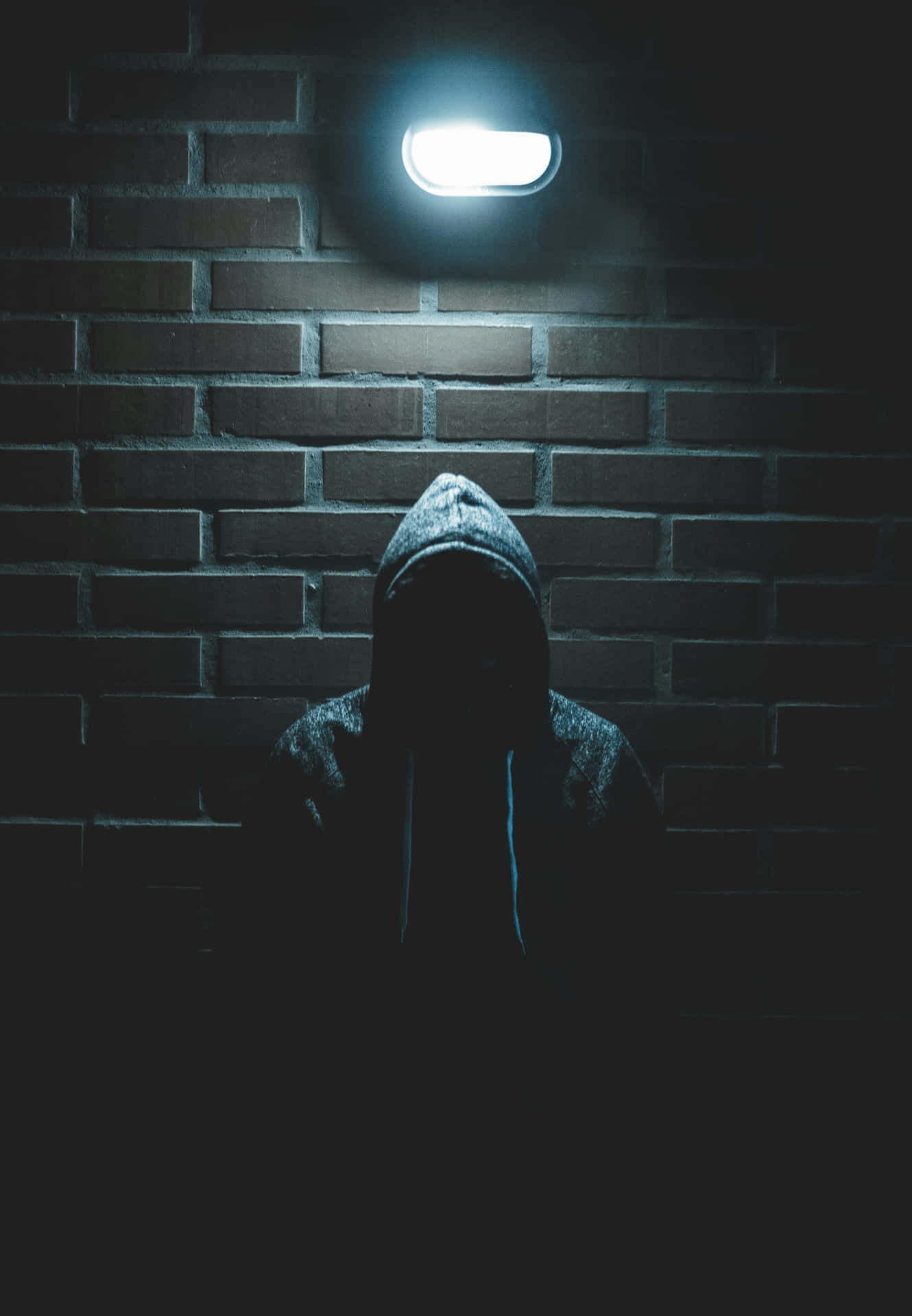  壁紙 — 一個戴著兜帽的身影站在磚牆前的陰影中，僅由頭頂上一盞燈照亮，散發出冰冷的藍色光芒。