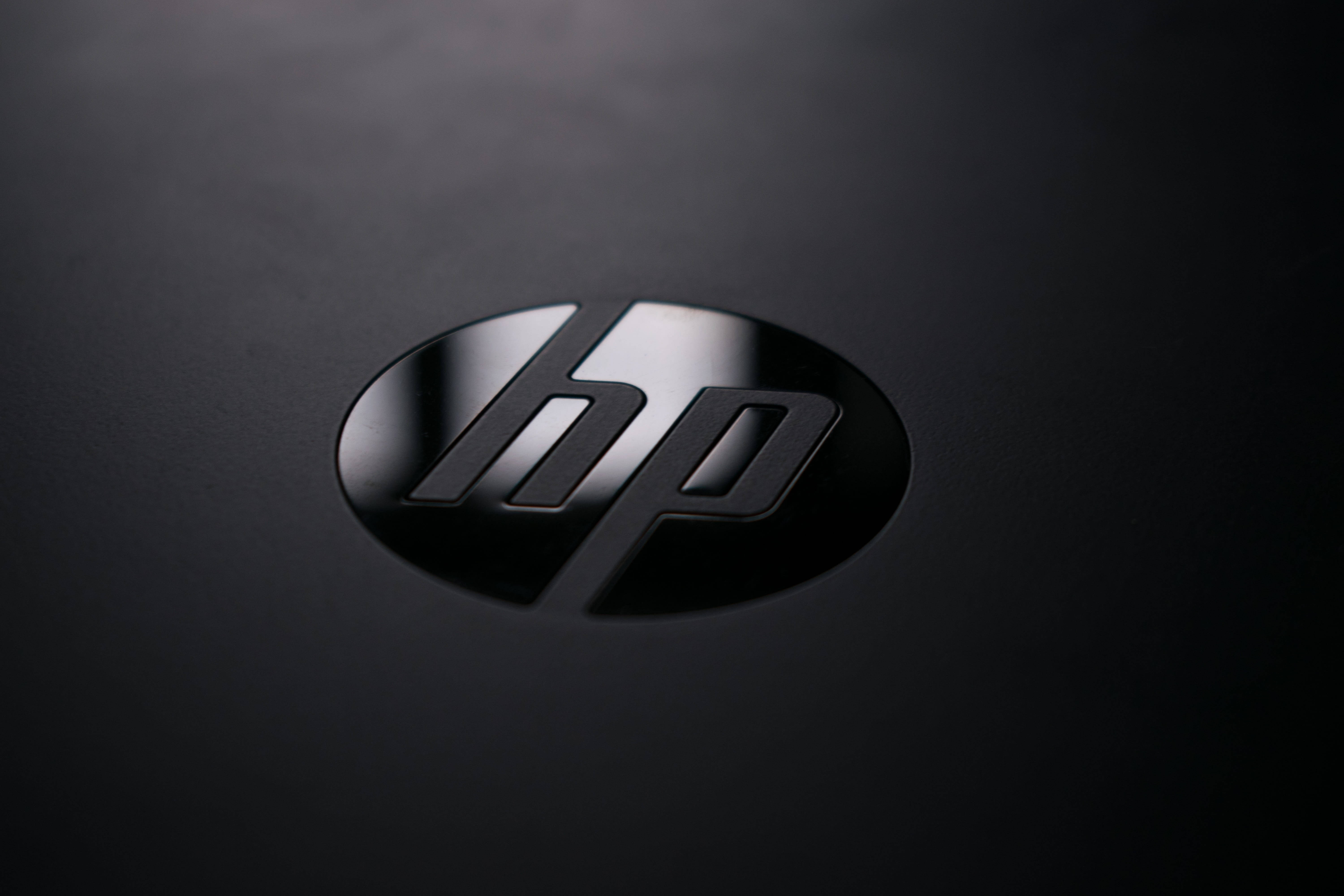 Hp Laptop 桌布 壁紙 — HP 標誌特寫，位於霧面黑色表面上，柔和光線突顯其金屬光澤。