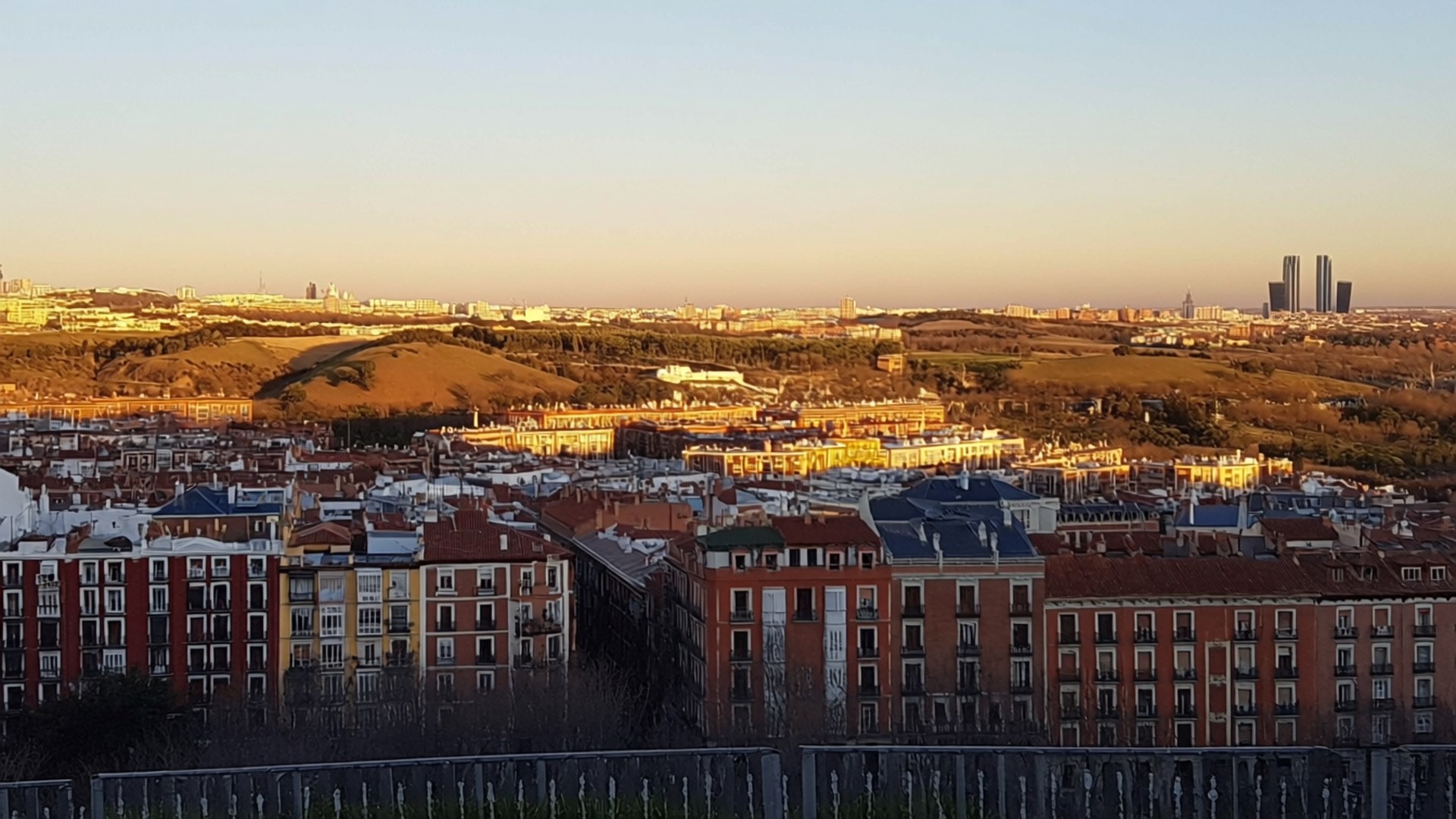 Madrid 桌布 壁紙 — 黃金時段的陽光沐浴著廣闊的城市景觀，陶土屋頂和遠處的摩天大樓映襯著柔和的漸變天空。