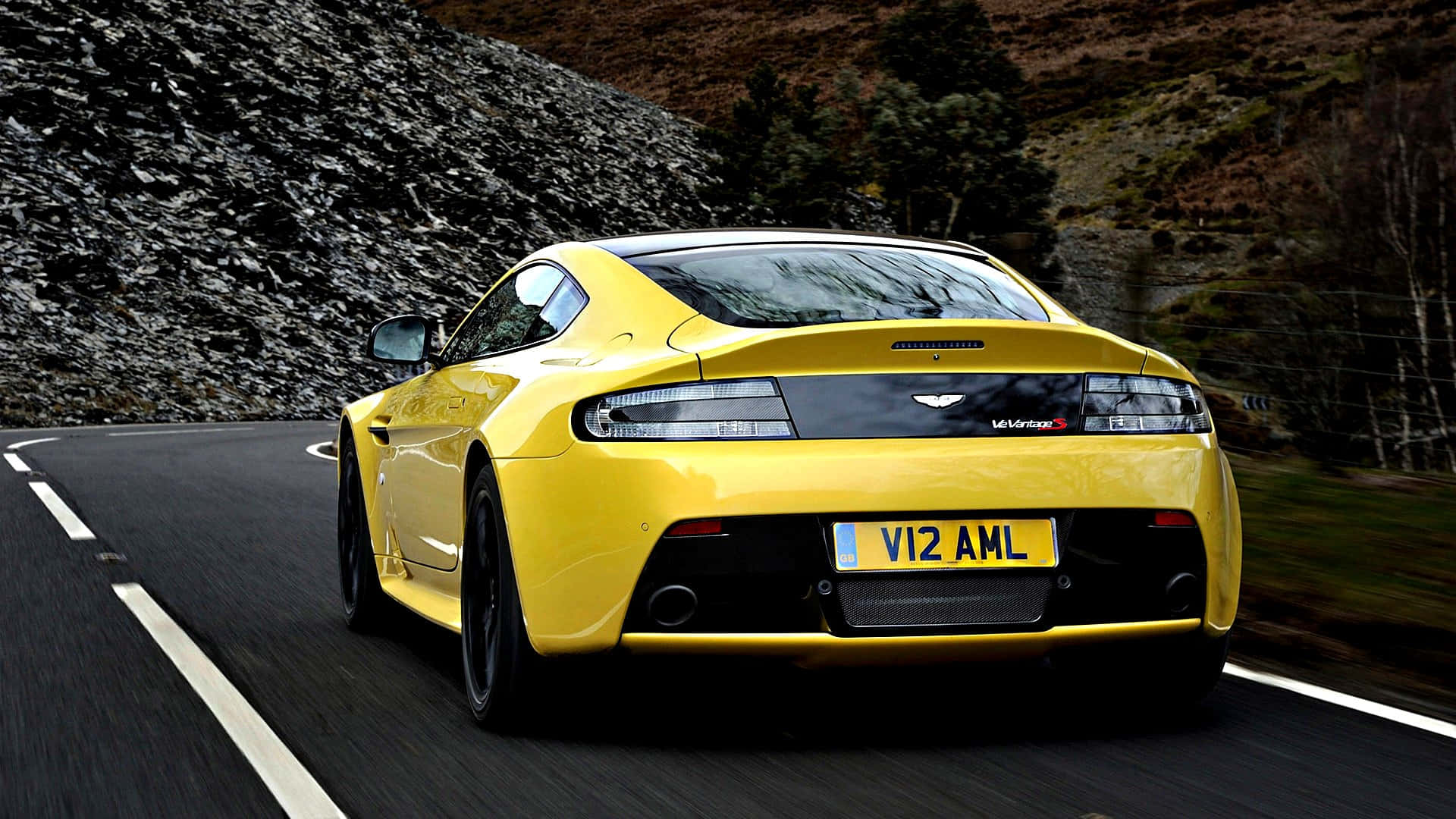 Aston Martin Vantage 桌布 壁紙 — 一輛鮮黃色的Aston Martin V12 Vantage在蜿蜒的山路上疾馳，帶有模糊的動態和岩石地形。