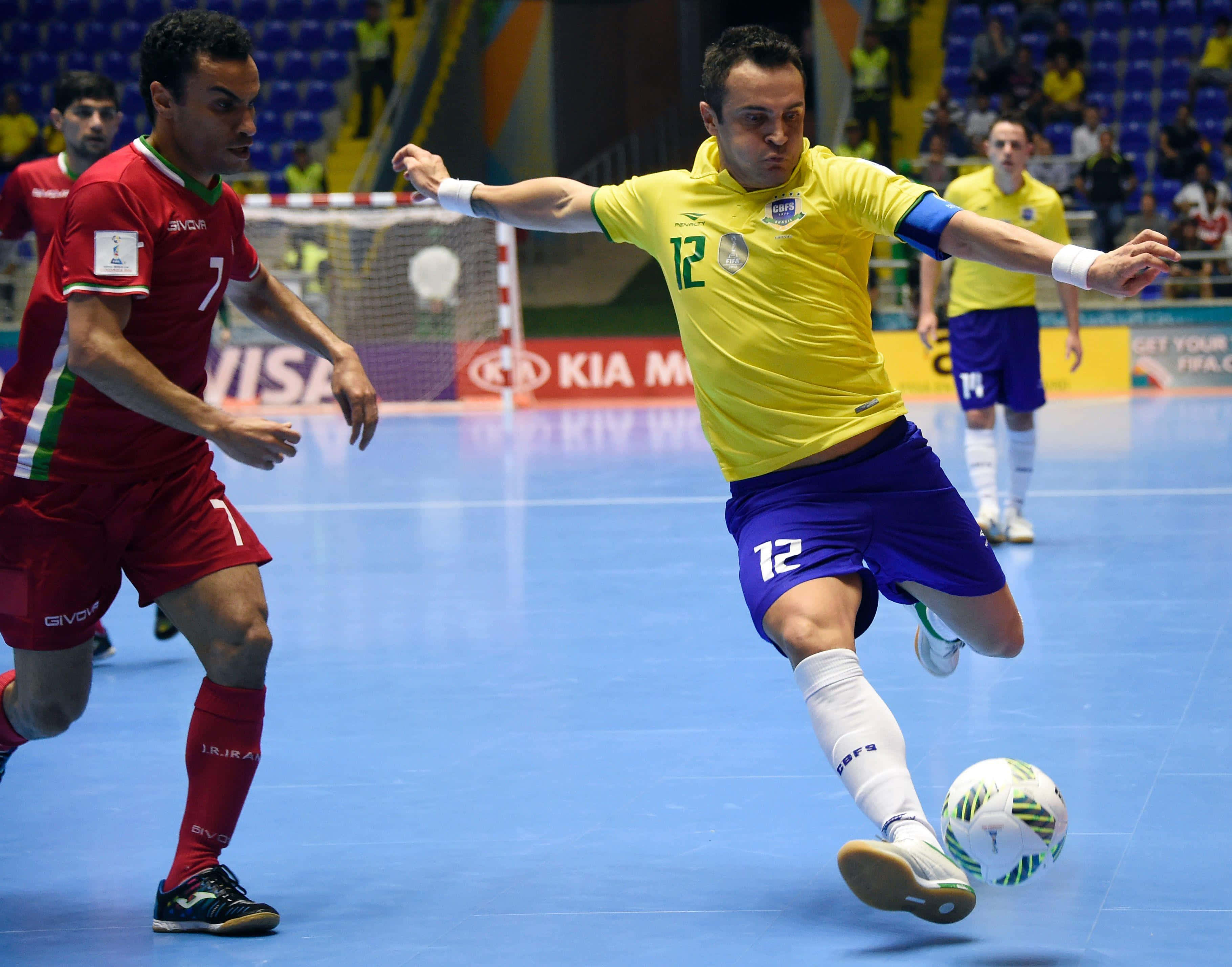 Indoor Soccer 桌布 壁紙 — 一名身穿黃藍球衣的室內足球員踢球，而一名紅衣對手在室內場地防守。