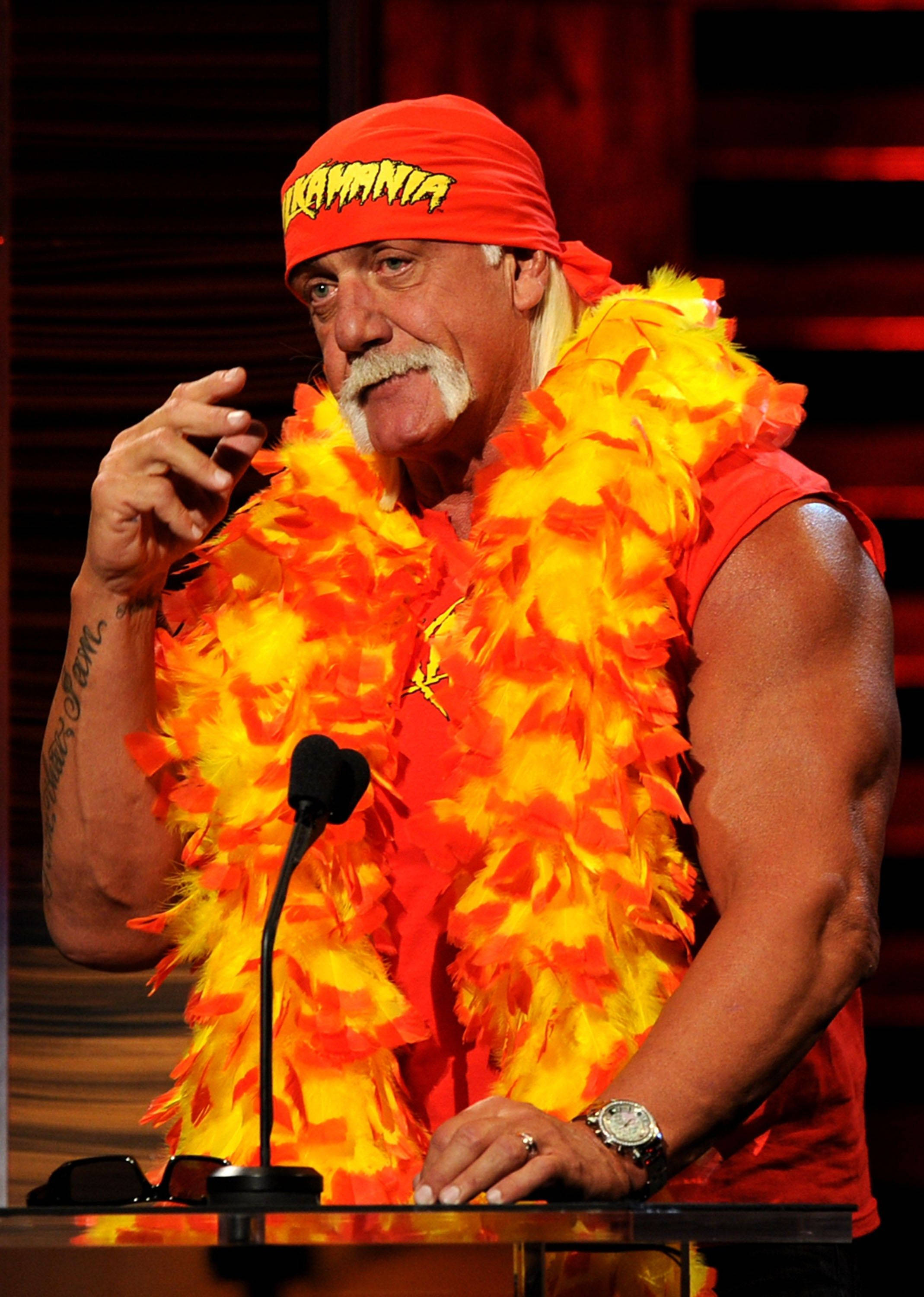 Hulk Hogan 桌布 壁紙 — Hulk Hogan 身穿標誌性的橙色和黃色羽毛服裝，站在舞台燈光下的講台上。