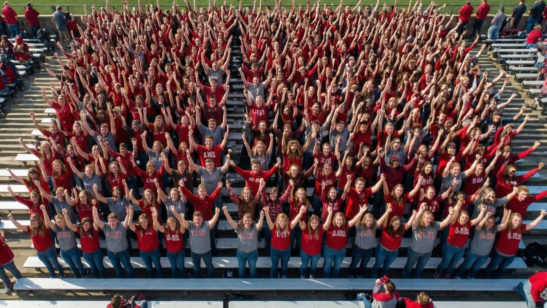 O-H-I-O Cheer 壁紙 — 數百名身穿紅色和灰色襯衫的學生在明亮的陽光下，從體育場看台上舉起手臂歡呼。