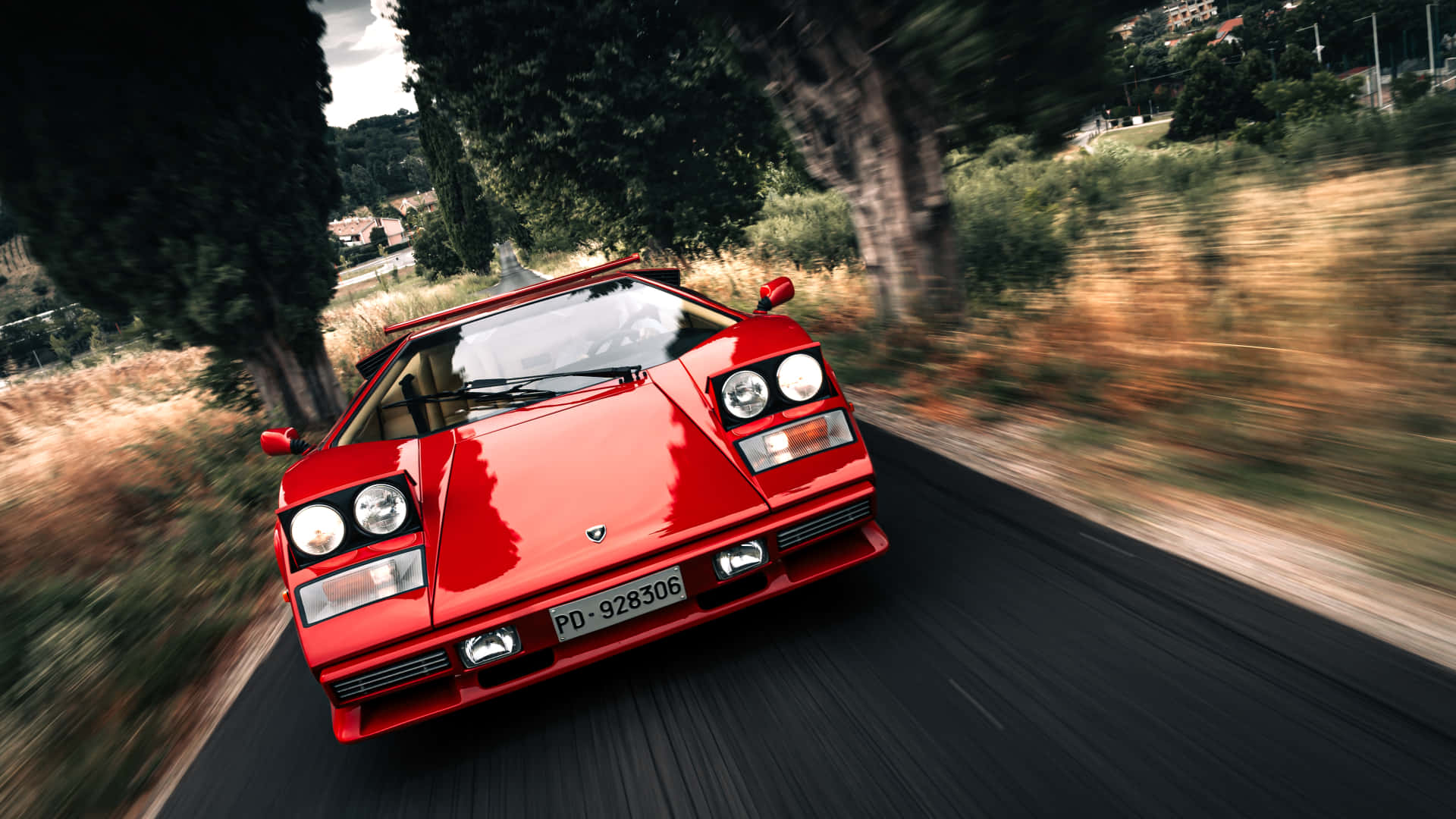 Lamborghini Countach 桌布 壁紙 — 一輛鮮紅色的藍寶堅尼Countach在晴朗天空下，沿著兩旁有高大樹木和金色草地的蜿蜒道路飛馳。