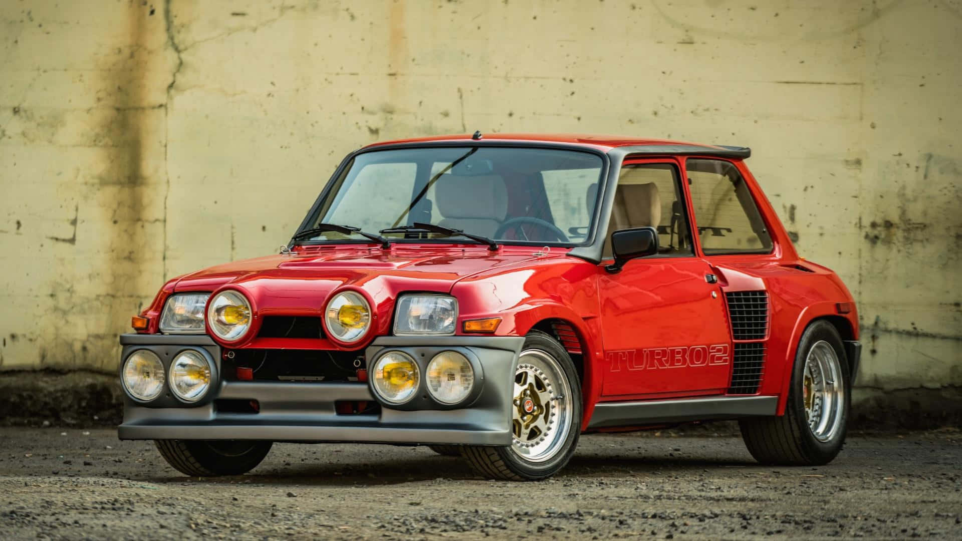 Renault 5 Turbo 桌布 壁紙 — 一輛鮮紅色的雷諾5 Turbo 2跑車，配備六個圓形頭燈和賽車條紋，自豪地停靠在風化的混凝土牆前。