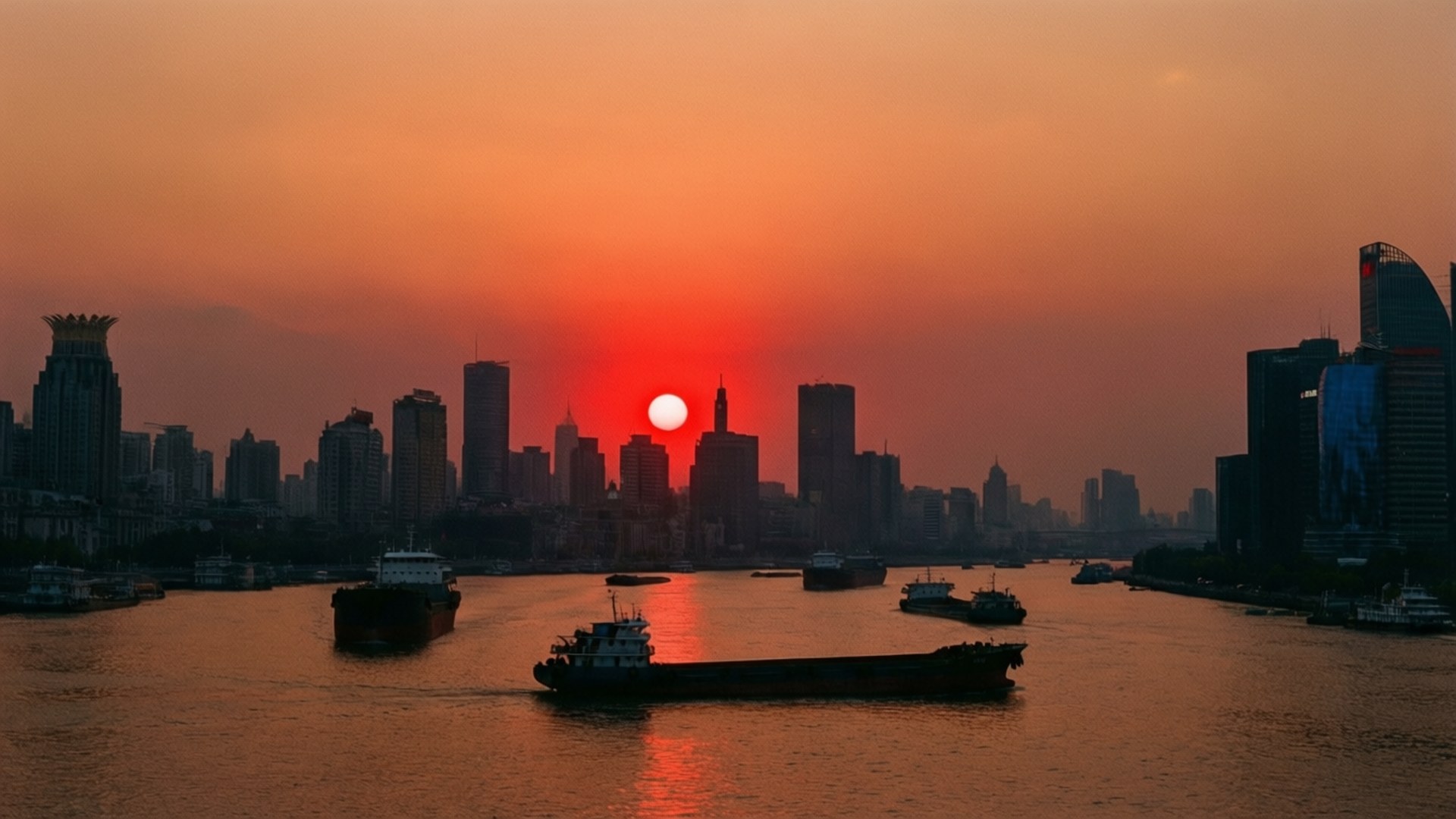 Sunset Over Huangpu 壁紙 — 火熱的橙色夕陽在現代城市天際線後落下，輪廓鮮明的貨船在反射著光芒的河面上滑行。