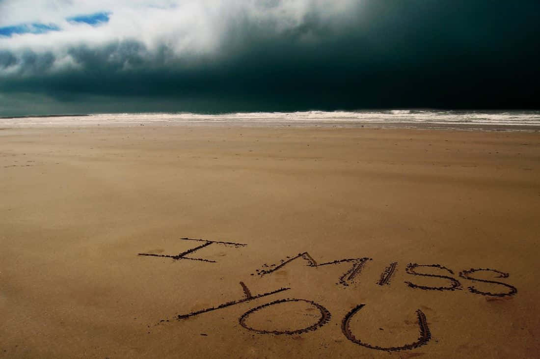 我想你 桌布 壁紙 — 「I MISS YOU」這幾個字刻在荒蕪海灘的濕沙上，上方籠罩著一片迫近的黑暗暴風雲。