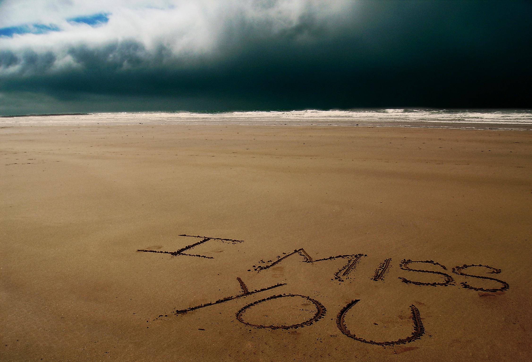 Missing 桌布 壁紙 — 「I MISS YOU」這幾個字刻在一個廣闊沙灘的濕沙上，上方是一片逼近的黑暗暴風雲。