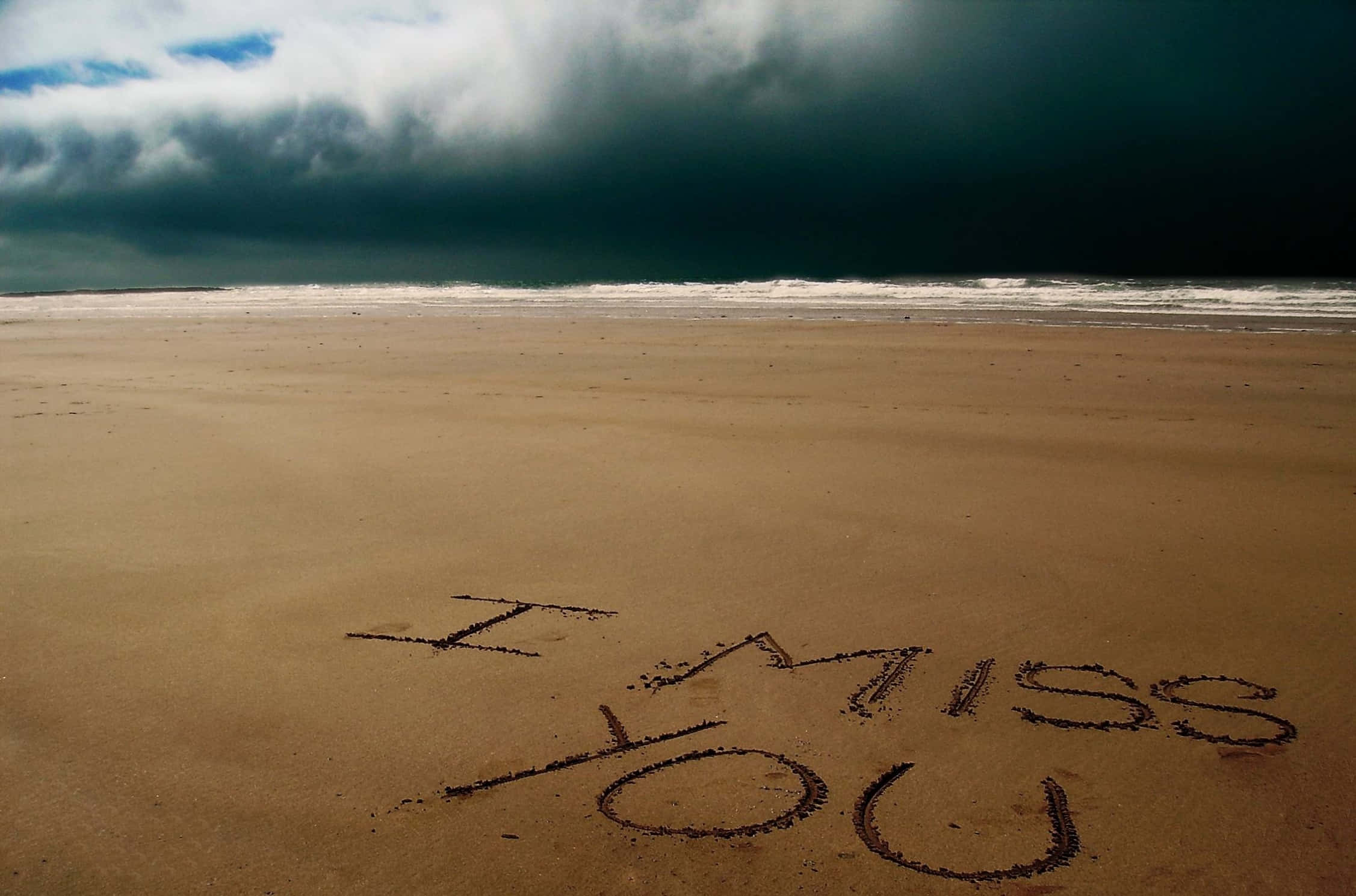 我想你 桌布 壁紙 — 「I MISS YOU」這幾個字刻在濕沙上，在空無一人的沙灘上，頭頂是一片逼近的黑暗暴風雲。