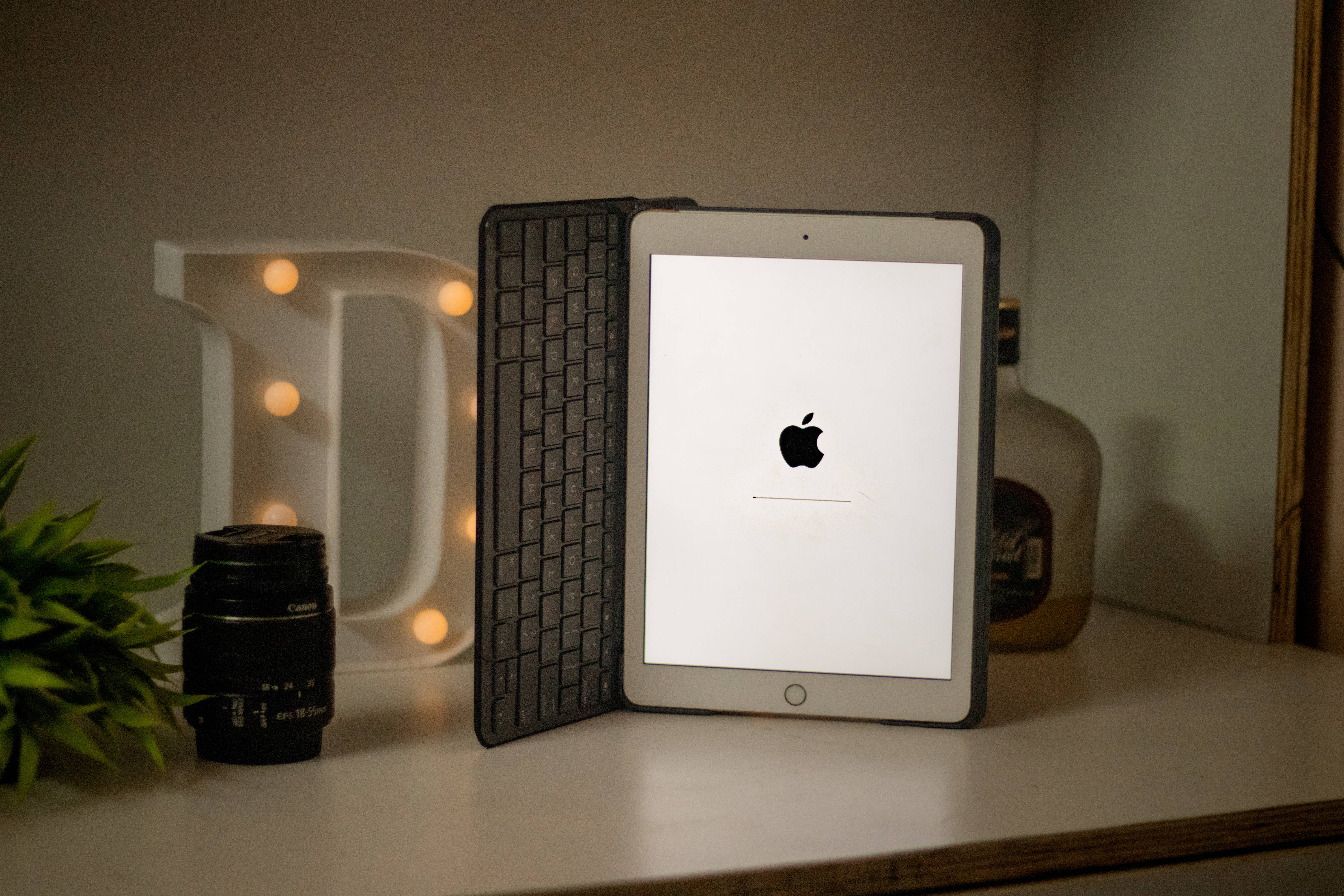 Mac 桌布 壁紙 — iPad 配備鍵盤保護套，在木架上顯示 Apple 標誌，旁邊有發光字母 D 相機鏡頭和植物。