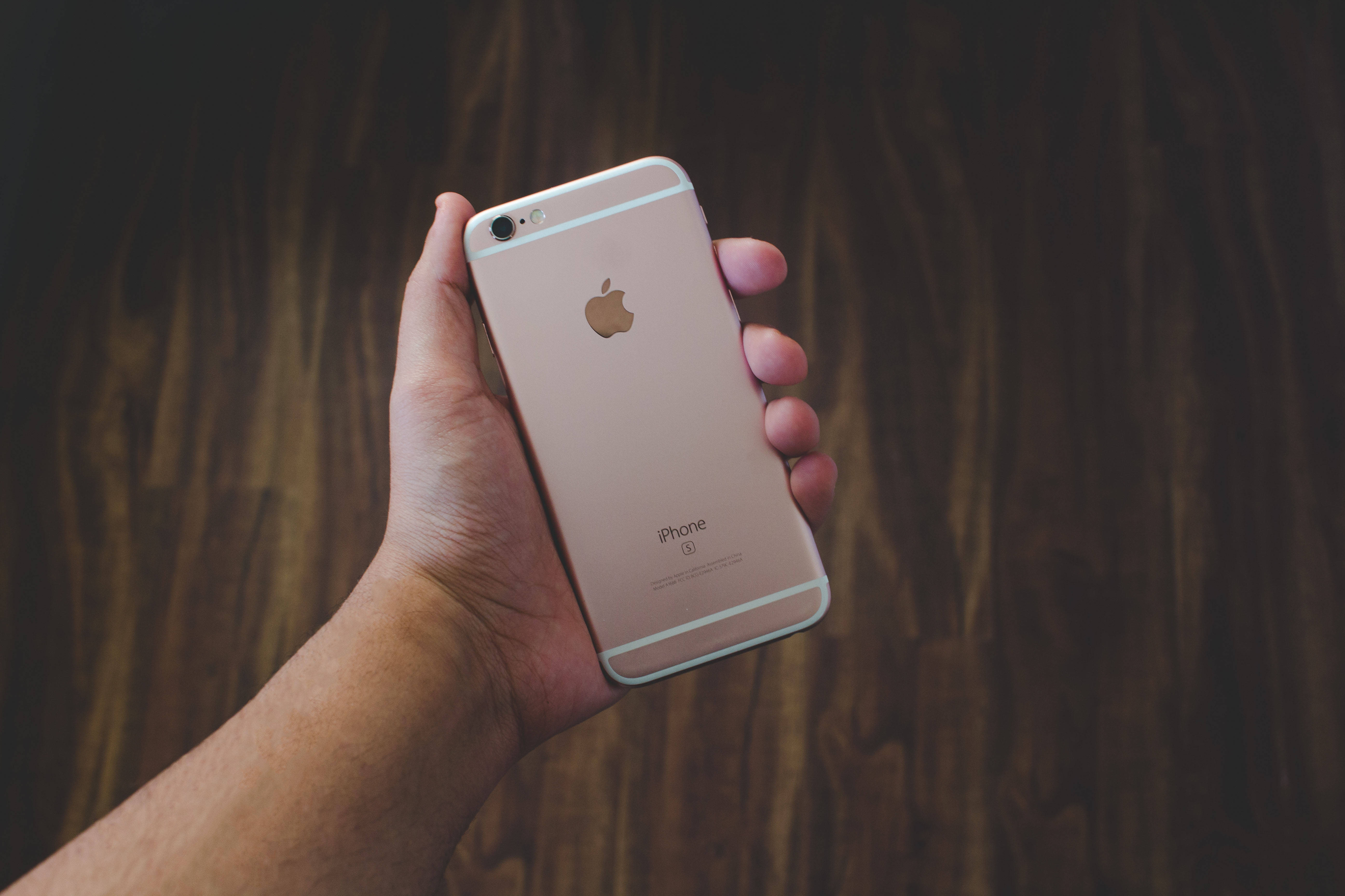 手持玫瑰金iPhone 6 壁紙 — 一隻手拿著玫瑰金iPhone 6s，背景為深色木材，光線柔和。