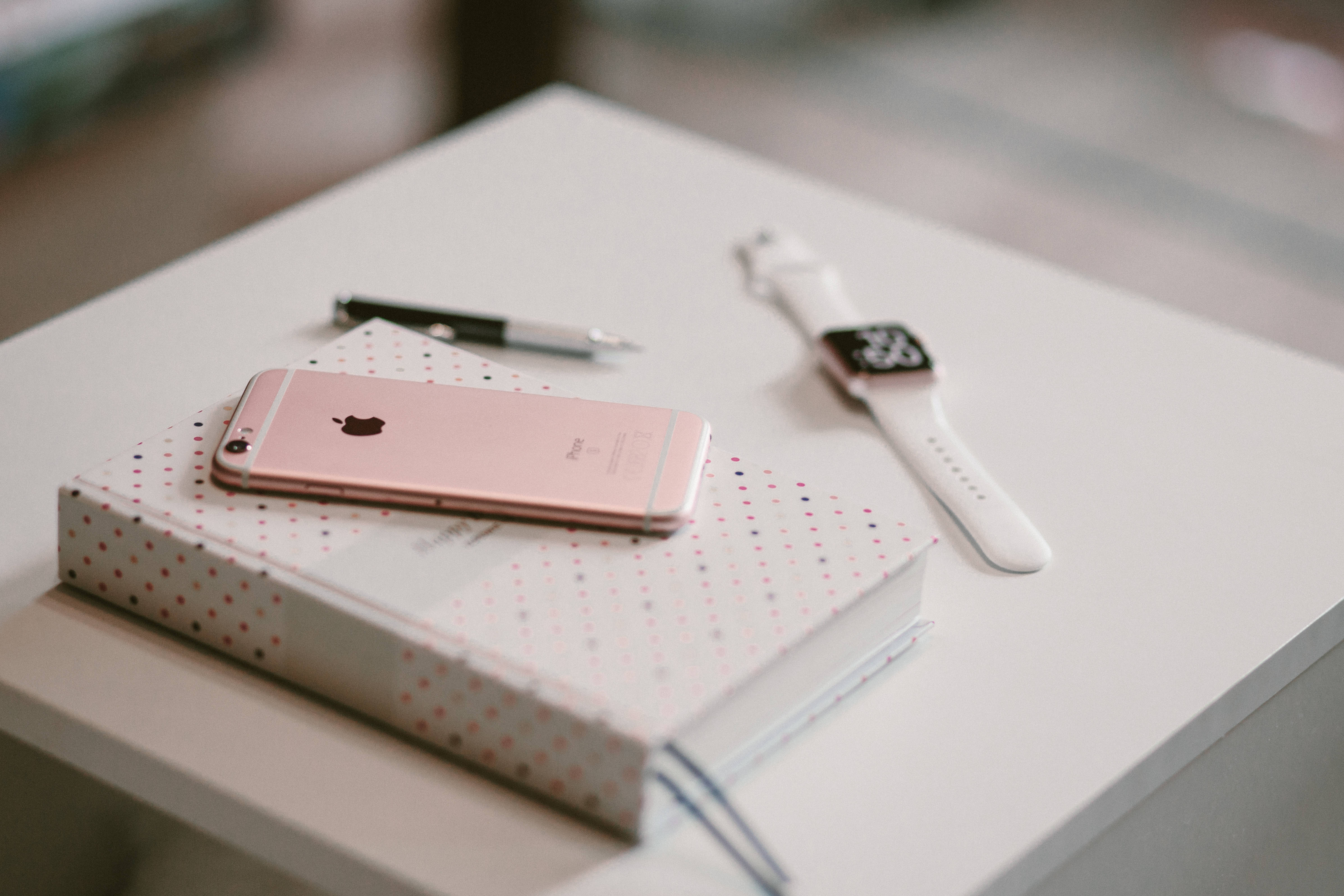 iPhone 6S 玫瑰金 壁紙 — 玫瑰金色 iPhone 放在圓點筆記本上，旁邊有白色 Apple Watch 和筆，置於簡約白色桌上。