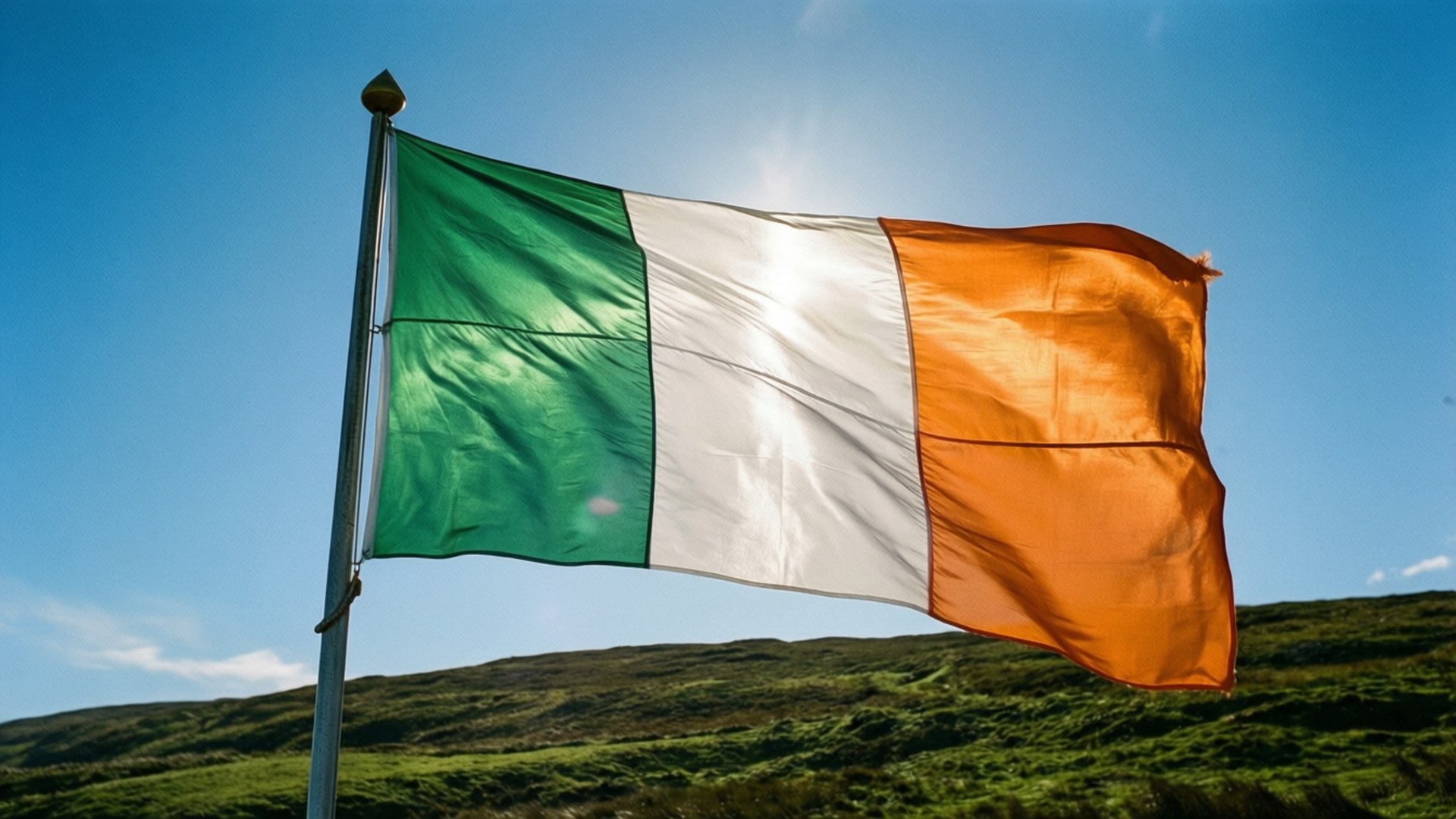 Irish Flag on Hill 壁紙 — 愛爾蘭國旗，帶有綠色、白色和橙色的垂直條紋，在晴朗的藍天下，在起伏的綠色山丘上自豪地飄揚。