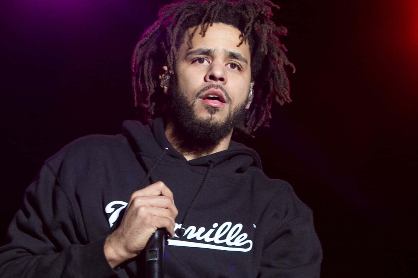  壁紙 — J. Cole 在戲劇性的紫色和紅色舞台燈光下，穿著黑色連帽衫在舞台上表演。