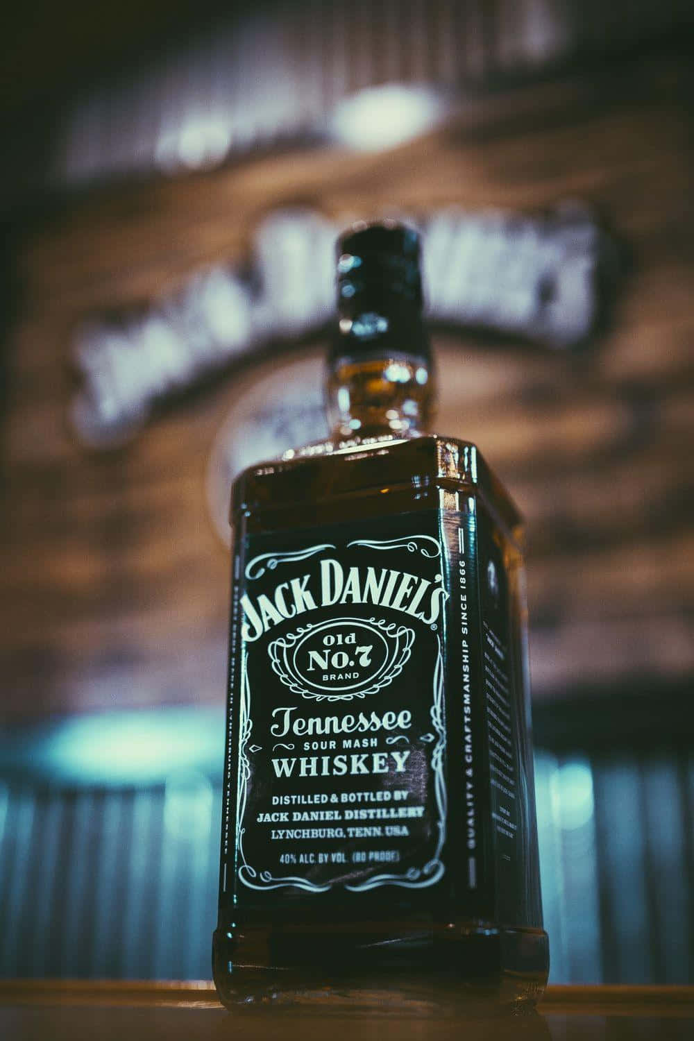 alcohol 桌布 壁紙 — Jack Daniel's 威士忌酒瓶在溫暖燈光下發光的特寫，背景是模糊的釀酒廠標誌。