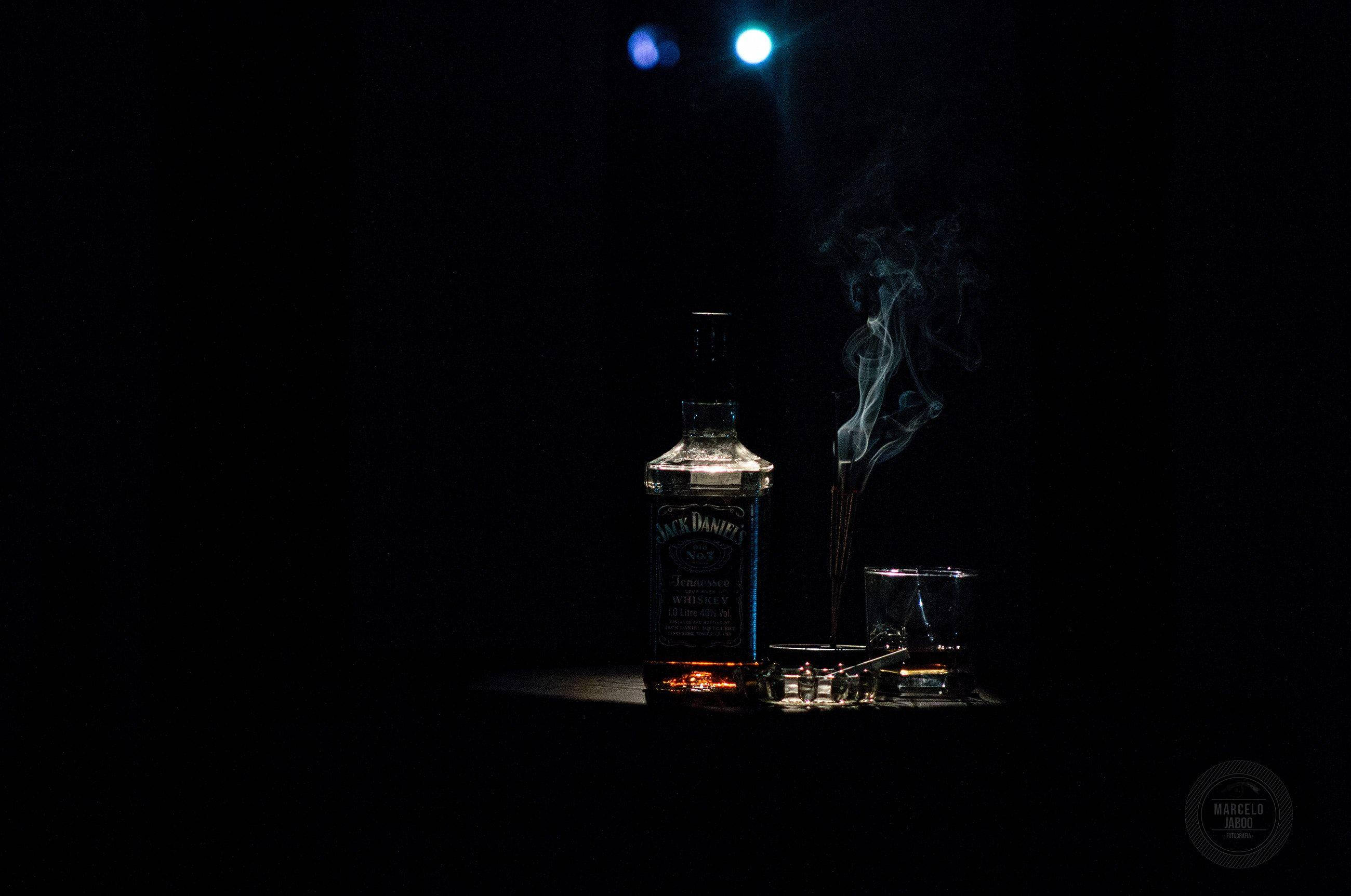 黑色 桌布 壁紙 — Jack Daniel's 酒瓶和酒杯在黑暗虛空中，於舞台燈光下閃耀，伴隨升起的煙霧。