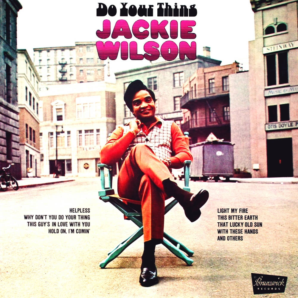 Singer 桌布 壁紙 — Jackie Wilson 自信地微笑，坐在導演椅上，身處陽光明媚的城市街道，周圍有復古建築和專輯文字。