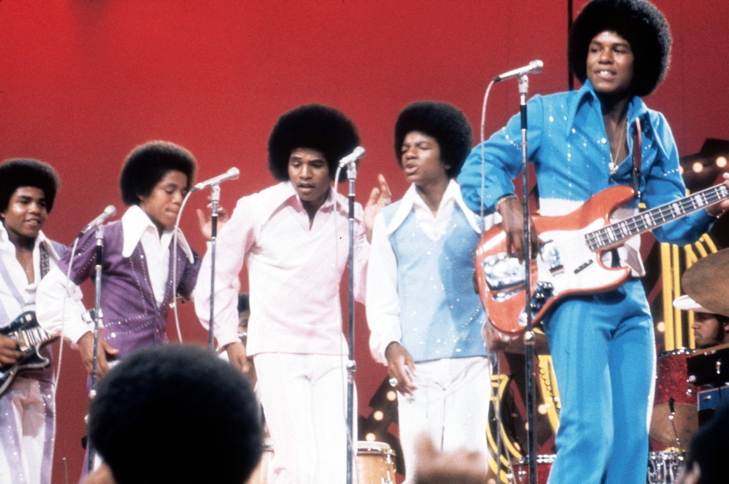 麥可·傑克森 桌布 壁紙 — Jackson Five 在舞台上穿著閃亮的70年代服裝，在鮮明的紅色背景前，手持麥克風和吉他表演。