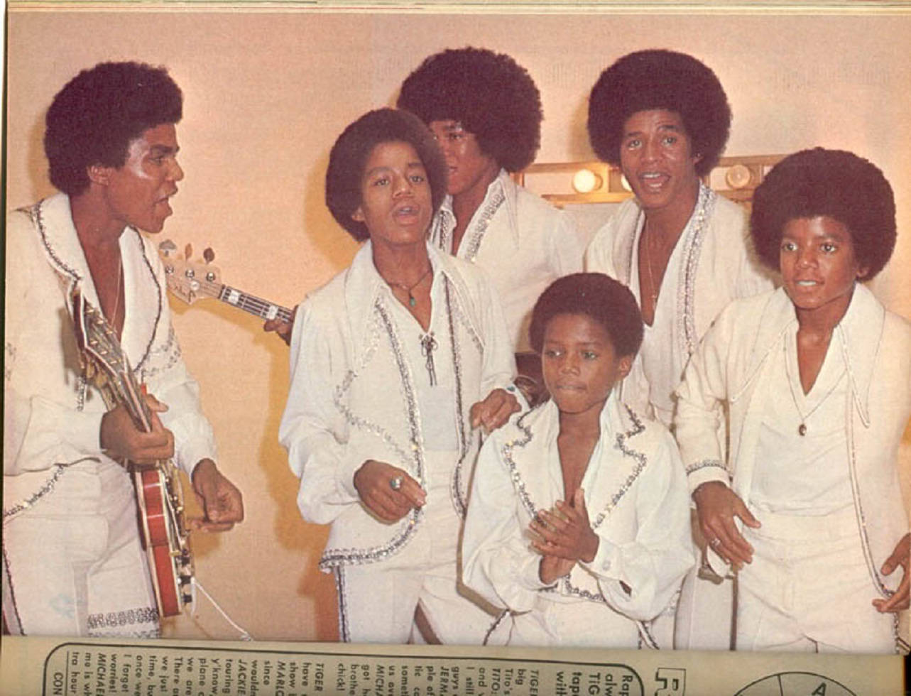 麥可·傑克森 桌布 壁紙 — Jackson Five 穿著相配的白色亮片服裝一起擺姿勢，其中一名成員拿著吉他，背景是暖色調的攝影棚。