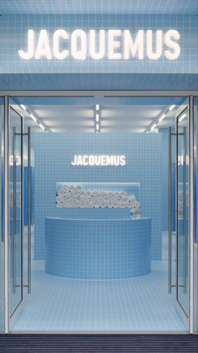 品牌 桌布 壁紙 — Jacquemus 店鋪內部採用極簡風格，配以藍色瓷磚、發光標誌和一個中央底座，上面放有捲起的毛巾。