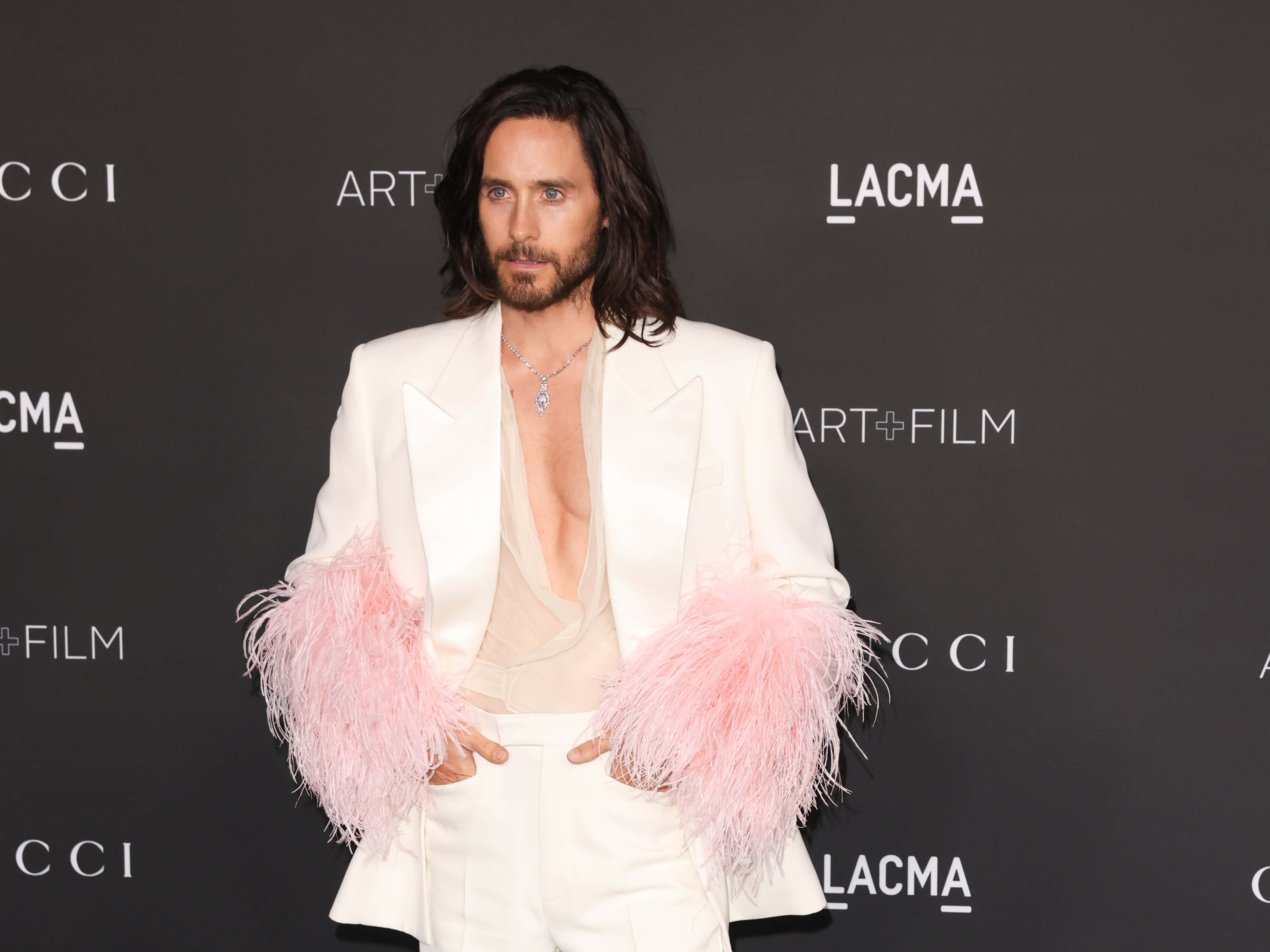 jared leto 桌布 壁紙 — Jared Leto 自信地穿著白色西裝，搭配粉紅色羽毛袖子，在黑色LACMA背景下擺姿勢。