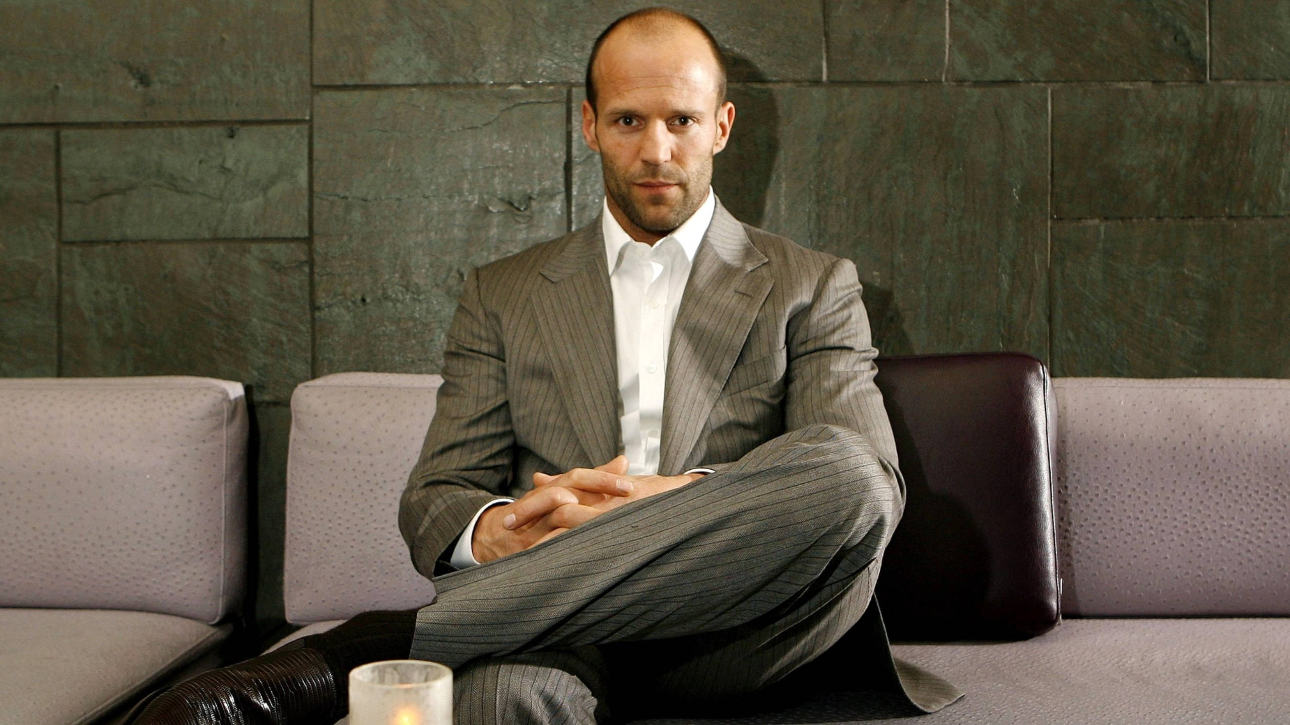 jason 桌布 壁紙 — Jason Statham 盤腿坐在現代沙發上，身穿細條紋西裝，背景是深色石牆，旁邊點著蠟燭。