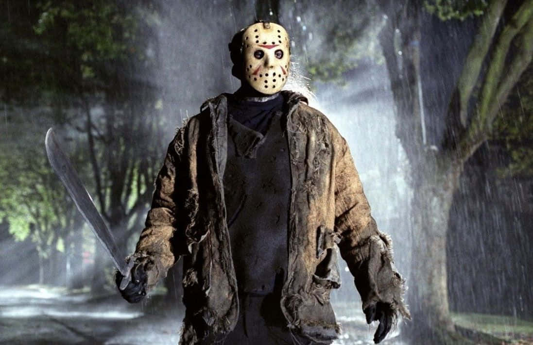 Creepy Halloween 桌布 壁紙 — Jason Voorhees 在夜晚的雨濕樹林小徑上威嚇地站立，手持砍刀，戴著他標誌性的曲棍球面具。
