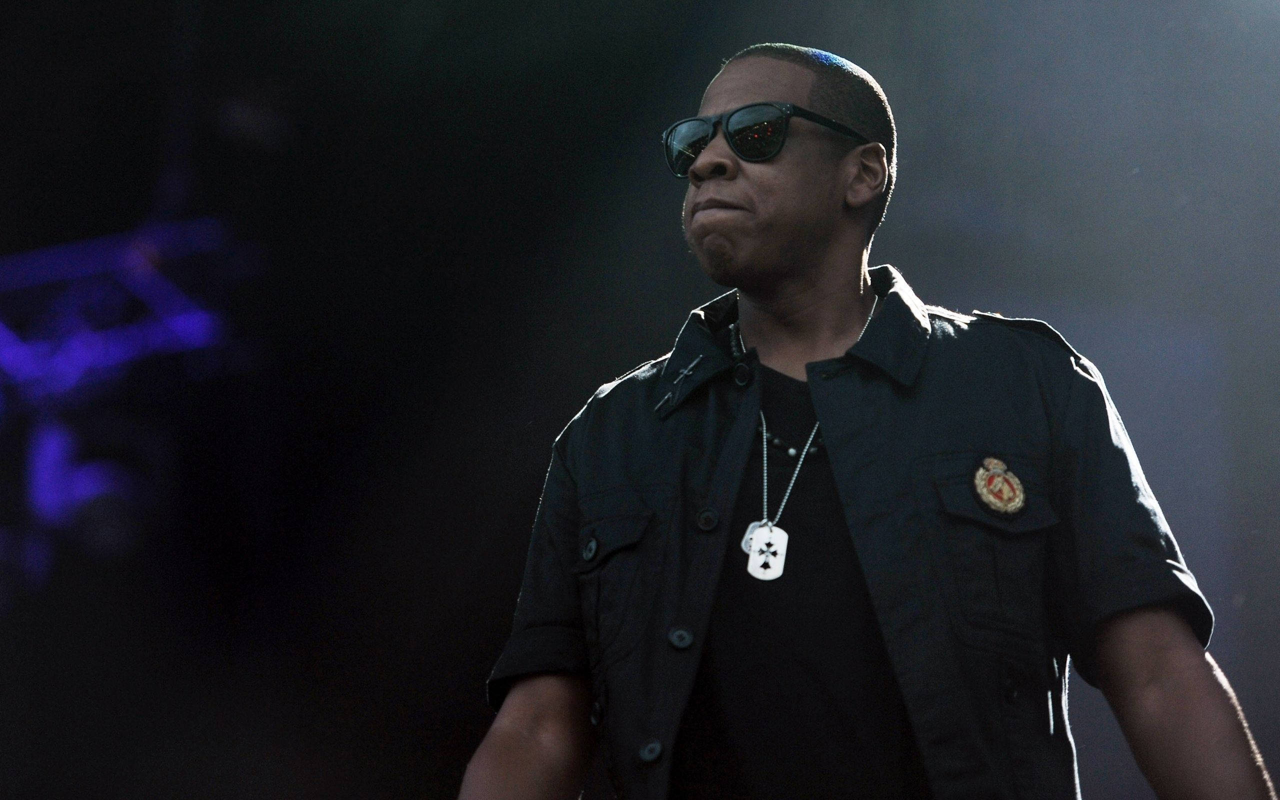 90s Rapper 桌布 壁紙 — JAY Z 自信地站在舞台上，戴著太陽眼鏡和穿著帶有狗牌的黑色襯衫，在戲劇性的紫色燈光下。