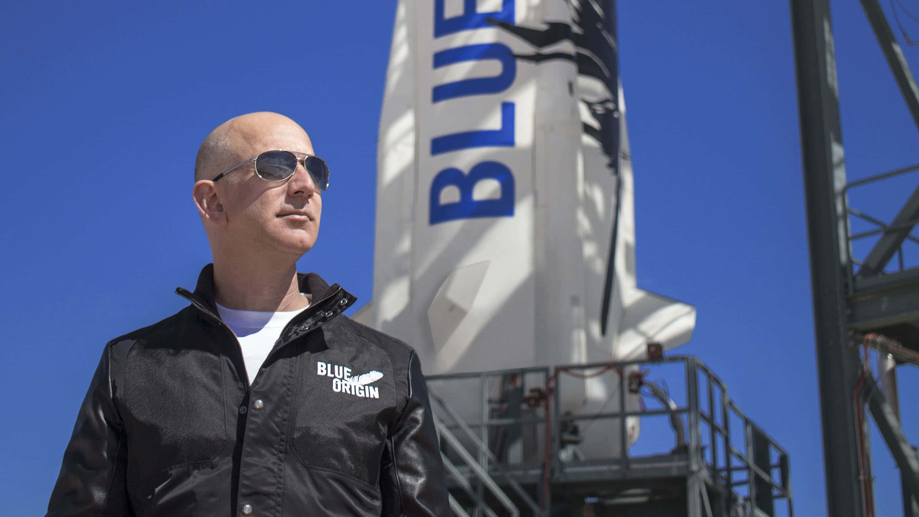 Jeff Bezos 桌布 壁紙 — Jeff Bezos 戴著太陽眼鏡和穿著 Blue Origin 外套，站在晴朗藍天下高聳的白色火箭前。