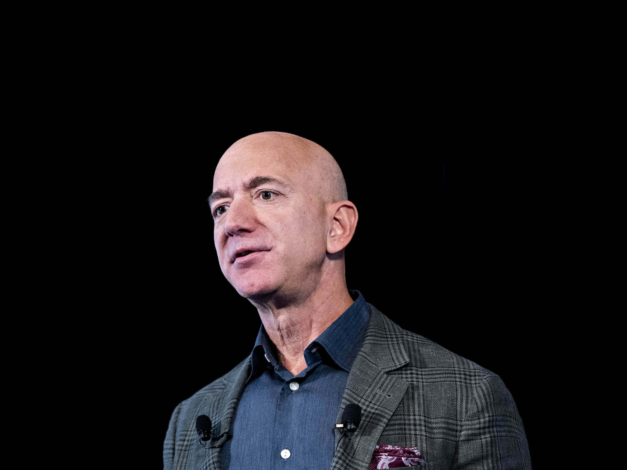Jeff Bezos 桌布 壁紙 — Jeff Bezos 身穿格紋西裝外套和藍色襯衫，站在純黑色背景前，在簡報時顯得若有所思。