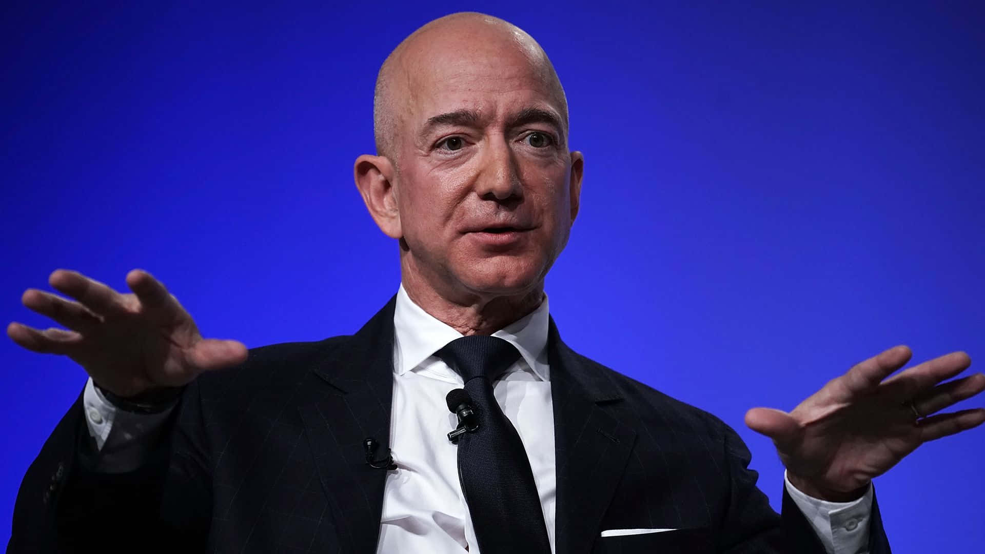 Jeff Bezos 桌布 壁紙 — Jeff Bezos 身穿深色西裝，在正式簡報中對著純藍色背景做出誇張的手勢。