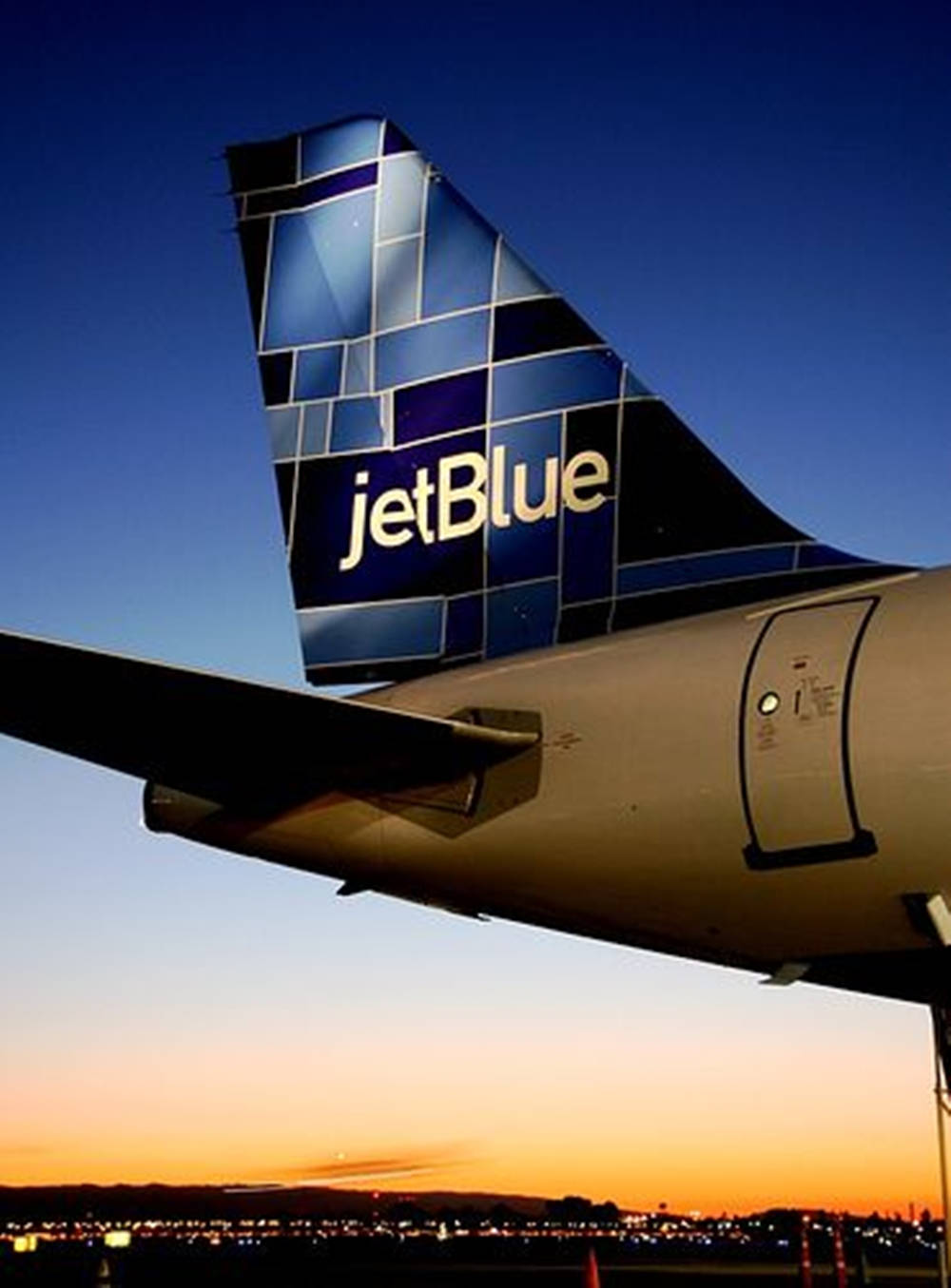 Plane 桌布 壁紙 — JetBlue 飛機尾部採用幾何藍色設計，在充滿活力的日落天空下，城市景觀中格外突出。
