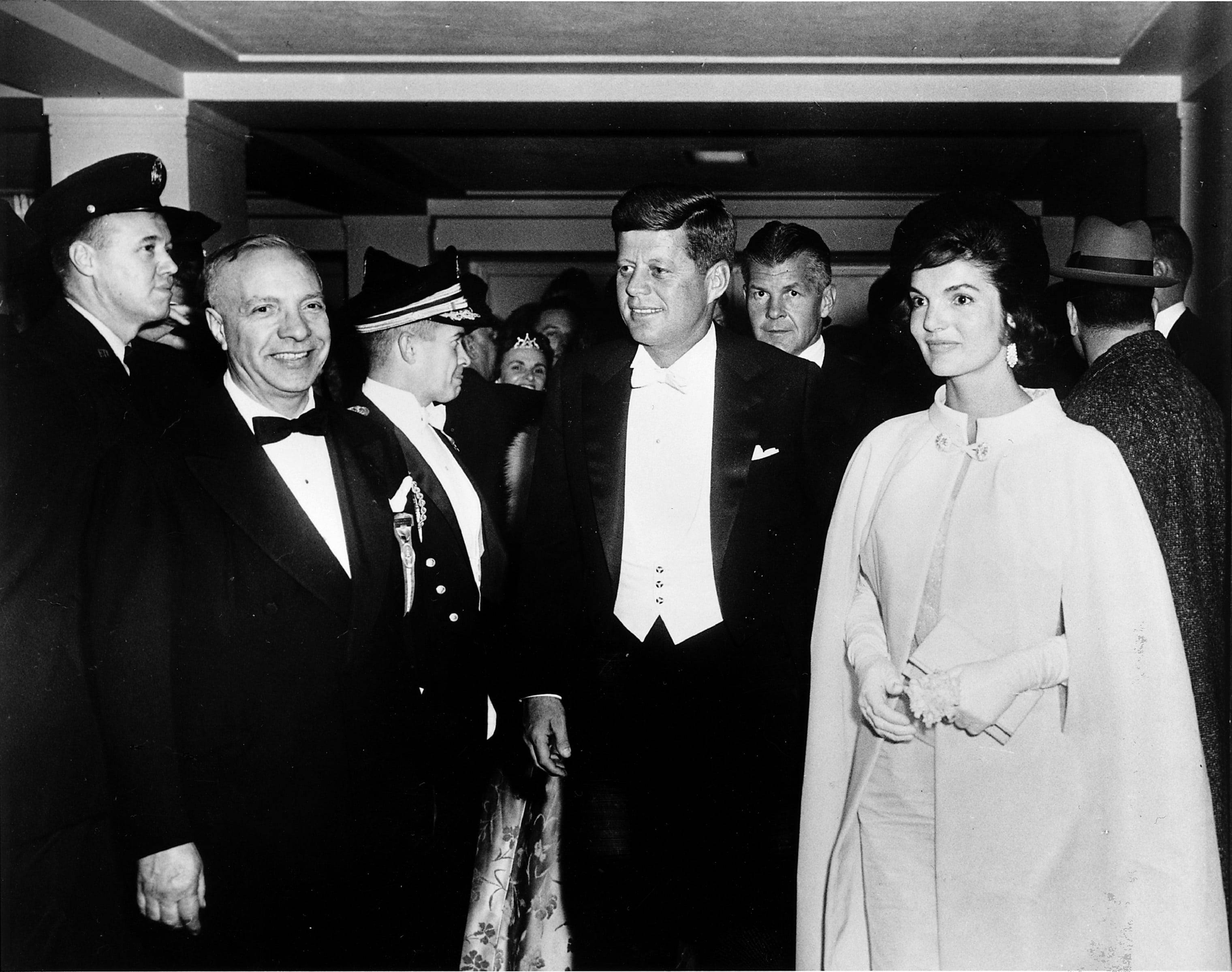 F 桌布 壁紙 — JFK 與賈桂琳·甘迺迪在正式室內聚會中微笑的黑白照片，周圍有穿制服的官員和賓客。