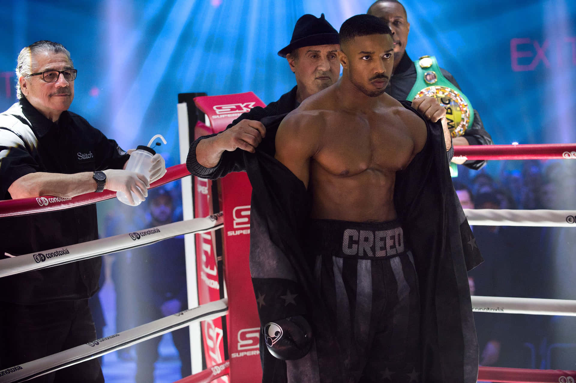  壁紙 — 一位肌肉發達的拳擊手穿著印有「CREED」的黑色短褲，站在燈光明亮的擂台上，教練正在脫下他的長袍，裁判在旁觀看。
