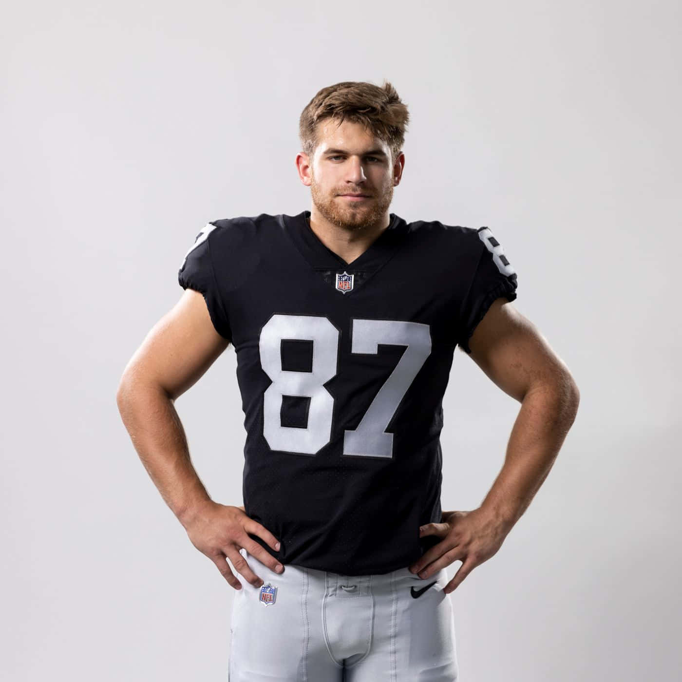  壁紙 — 一位肌肉發達的NFL球員身穿黑色球衣，號碼87，雙手叉腰，自信地站在純白色背景前。