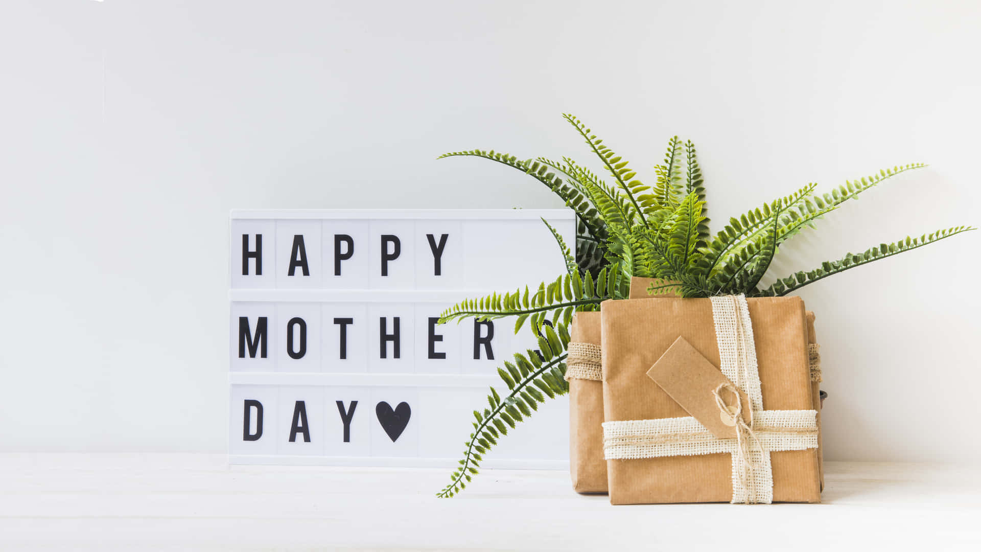  壁紙 — 一個簡約的白色架子上展示了一個「Happy Mother's Day」燈箱，旁邊有兩個用麻繩包裹的牛皮紙禮物和一棵茂盛的綠色蕨類植物。