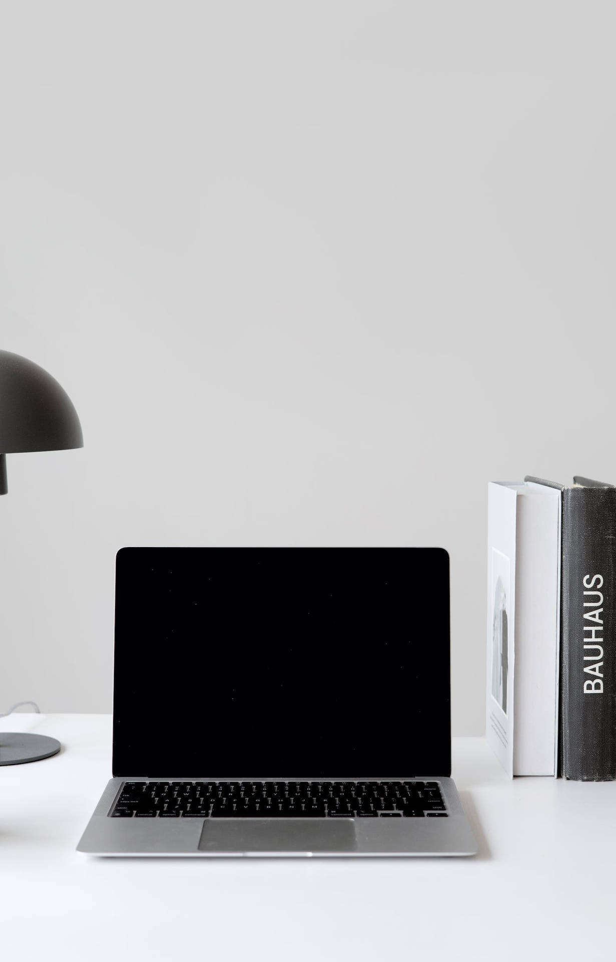 銀色筆記型電腦放在白色桌上，旁邊有黑色燈和包浩斯書籍。 — Macbook Pro 壁紙