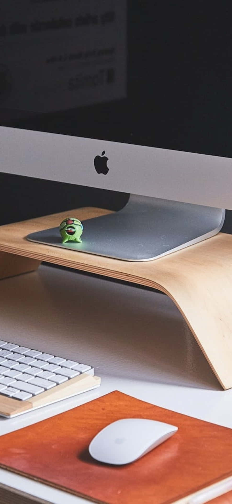 簡約電腦桌，配有 iMac、鍵盤、滑鼠和木製支架上的綠色小公仔。 — iPhone X 壁紙