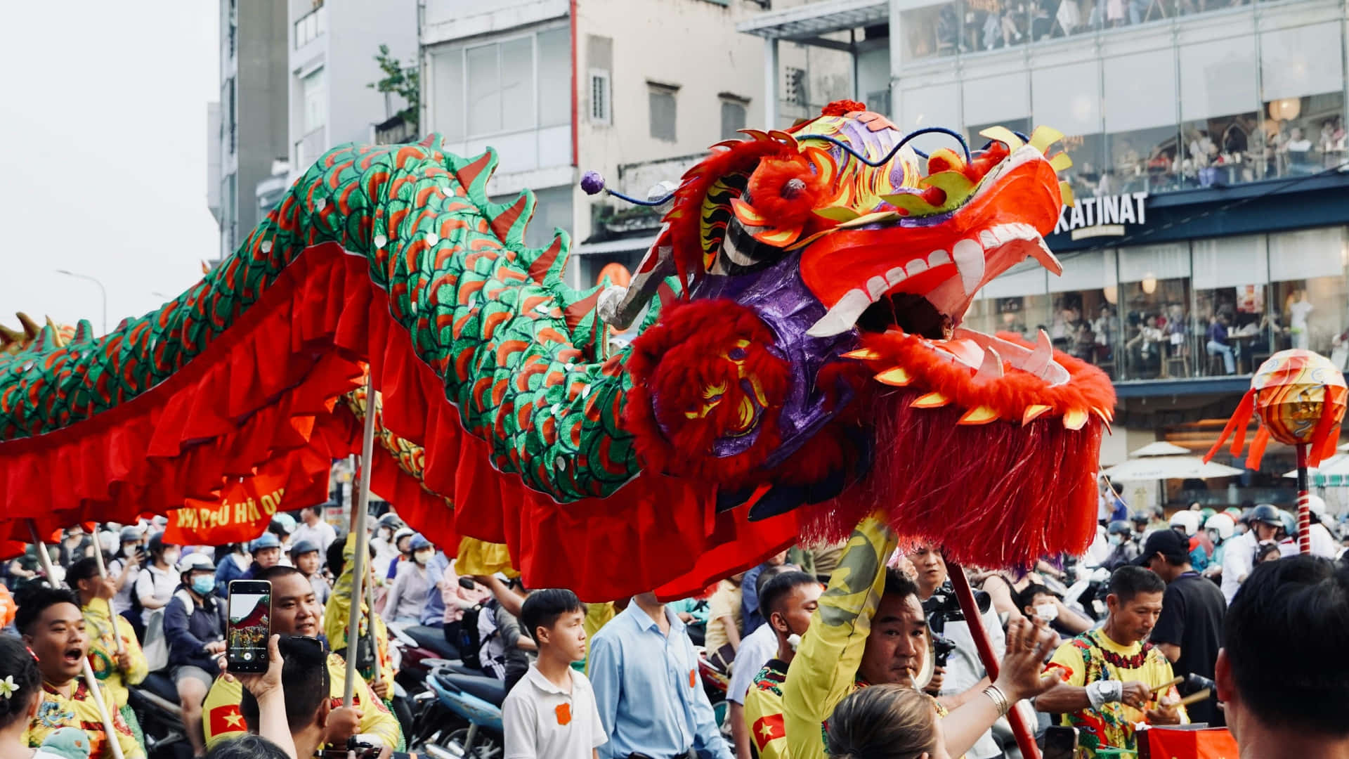  壁紙 — 節日遊行期間，表演者在擁擠的城市街道上進行充滿活力的紅色和綠色舞龍表演。