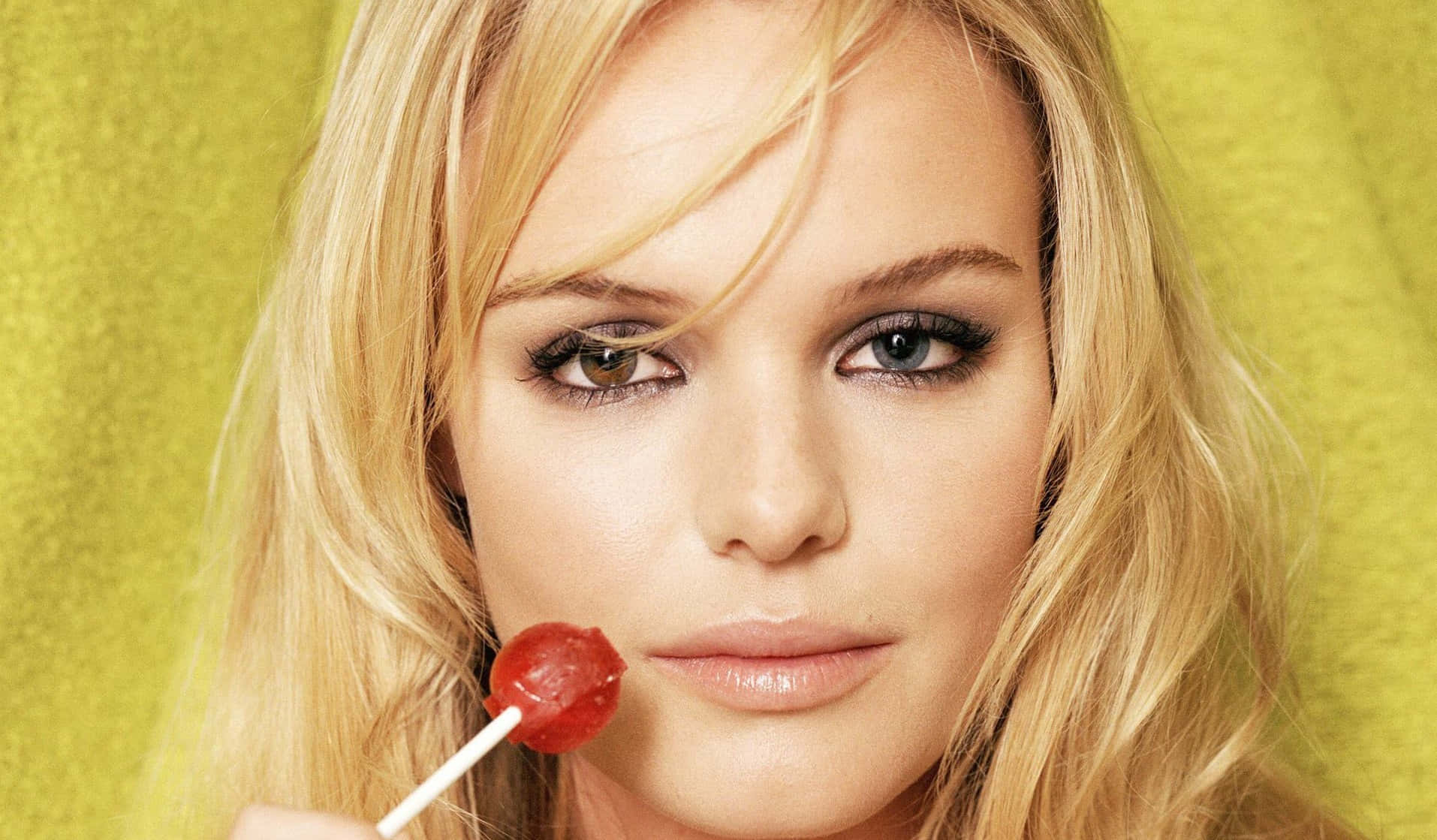 Kate Bosworth 桌布 壁紙 — 一位金髮碧眼的女人在充滿活力的黃綠色背景下，將紅色棒棒糖靠近嘴唇。
