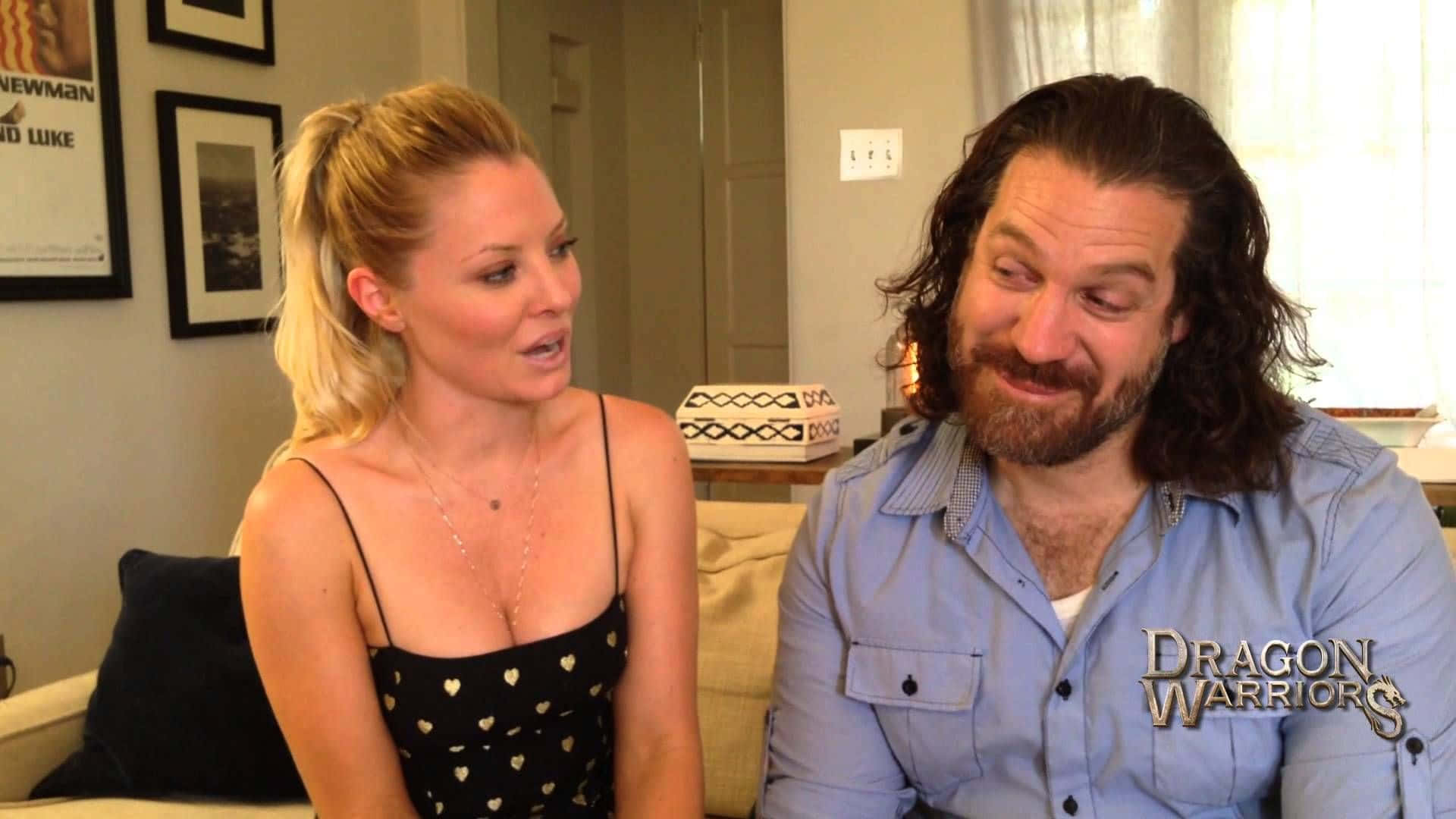 Kaitlin Doubleday 桌布 壁紙 — 一位金髮女子身穿黑色心形圖案上衣，一位留鬍鬚的男子身穿藍色襯衫，坐在沙發上，在燈光溫暖的客廳中交談。