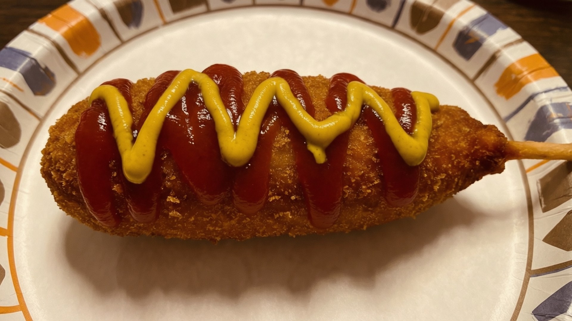 Corn Dog 桌布 壁紙 — 金黃色的玉米熱狗，上面有鋸齒狀的番茄醬和芥末，放在色彩繽紛的紙盤上。