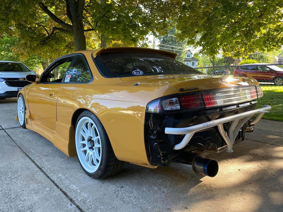  壁紙 — 一輛金黃色的日產S14跑車，配備白色輪圈和裸露的排氣管，停放在綠樹成蔭的混凝土車道上。