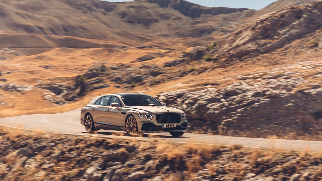 Bentley Mulsanne 桌布 壁紙 — 一輛金色賓利飛馳在明亮的日光下，沿著蜿蜒的山路穿過乾燥的金色山丘。