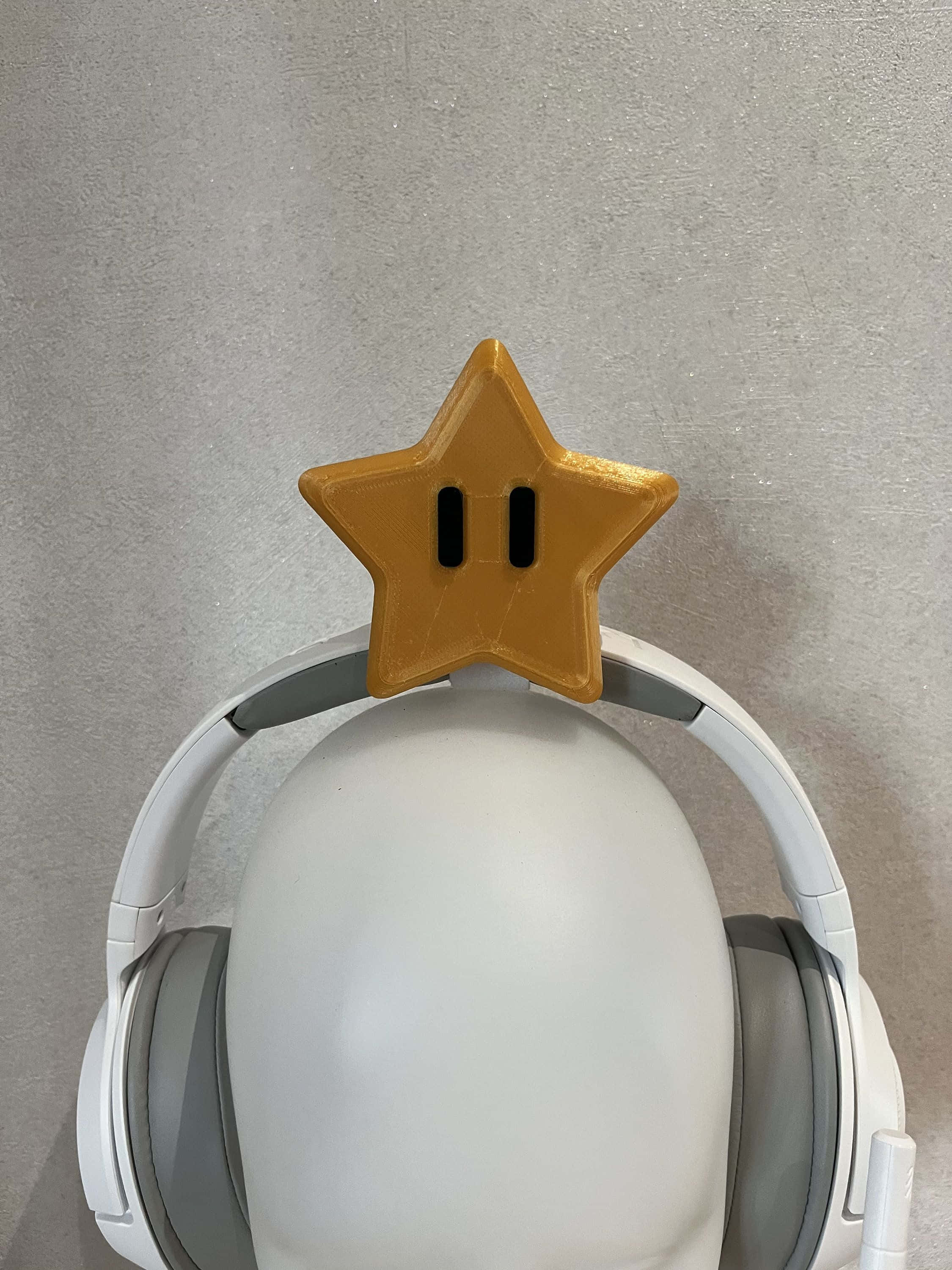 Gaming Accessories 桌布 壁紙 — 一顆金色的超級瑪利歐星星放在灰色牆壁前假人頭上的白色頭戴式耳機上。