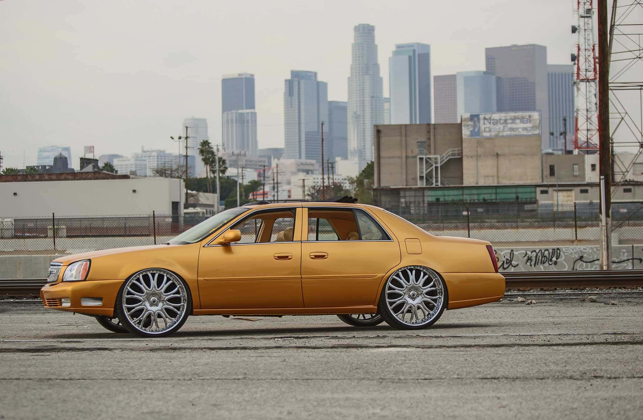Cadillac Deville 桌布 壁紙 — 一輛金色訂製凱迪拉克轎車，配備大型鍍鉻輪圈，停泊在俯瞰迷濛城市天際線的都市屋頂上。