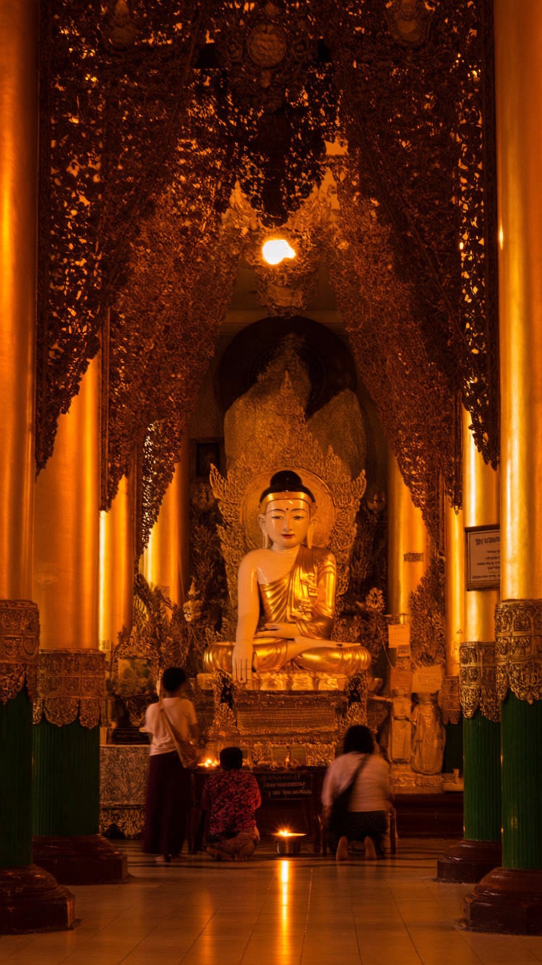 Inside Shwedagon Pagoda 壁紙 — 金色佛像在裝飾華麗的華蓋下靜坐，信徒們在溫暖的琥珀色燈光下跪拜祈禱。
